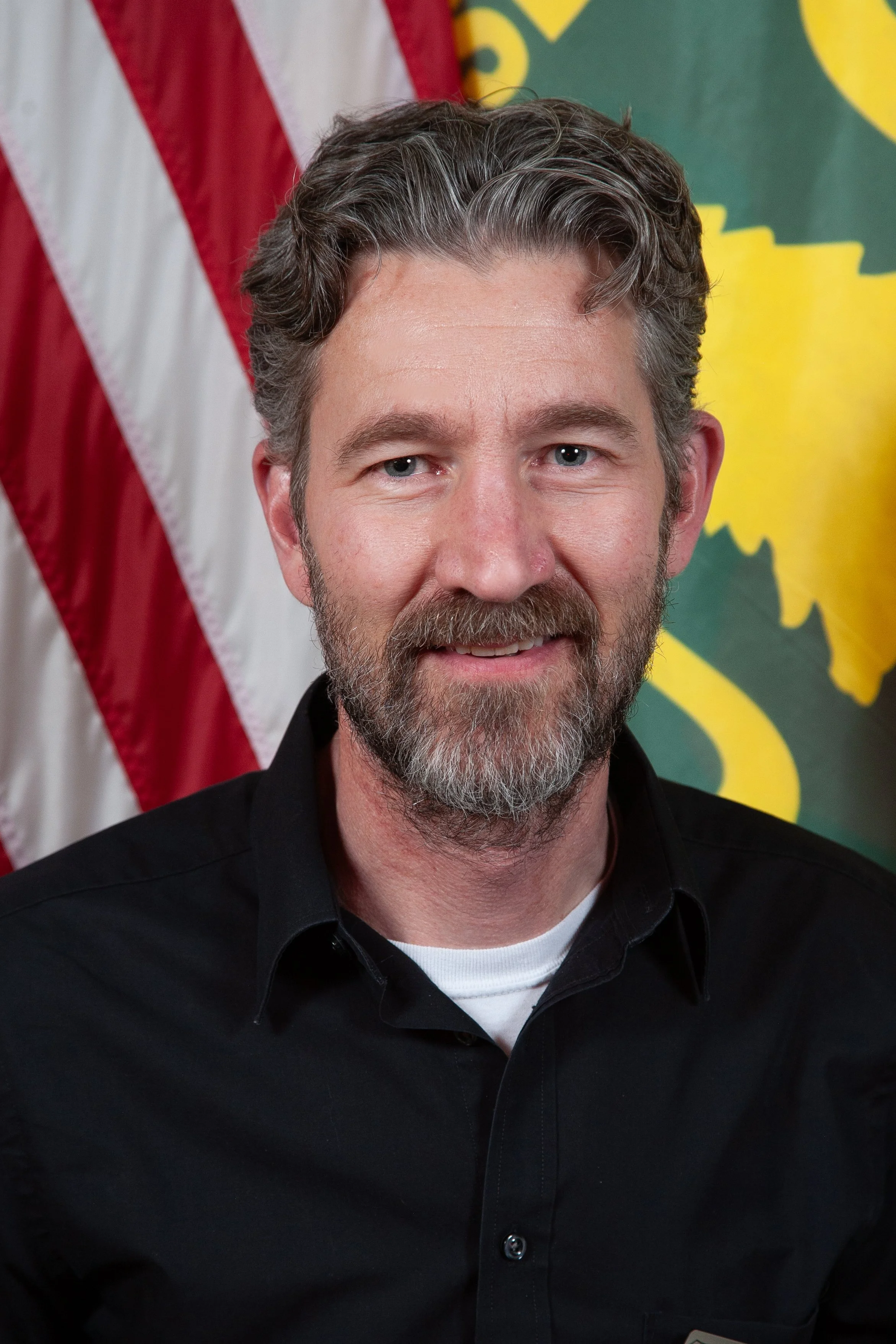 Close-up of a middle-aged man with gray and brown hair and a beard, wearing a black shirt. In the background, parts of the American flag and a green and yellow emblem are visible.