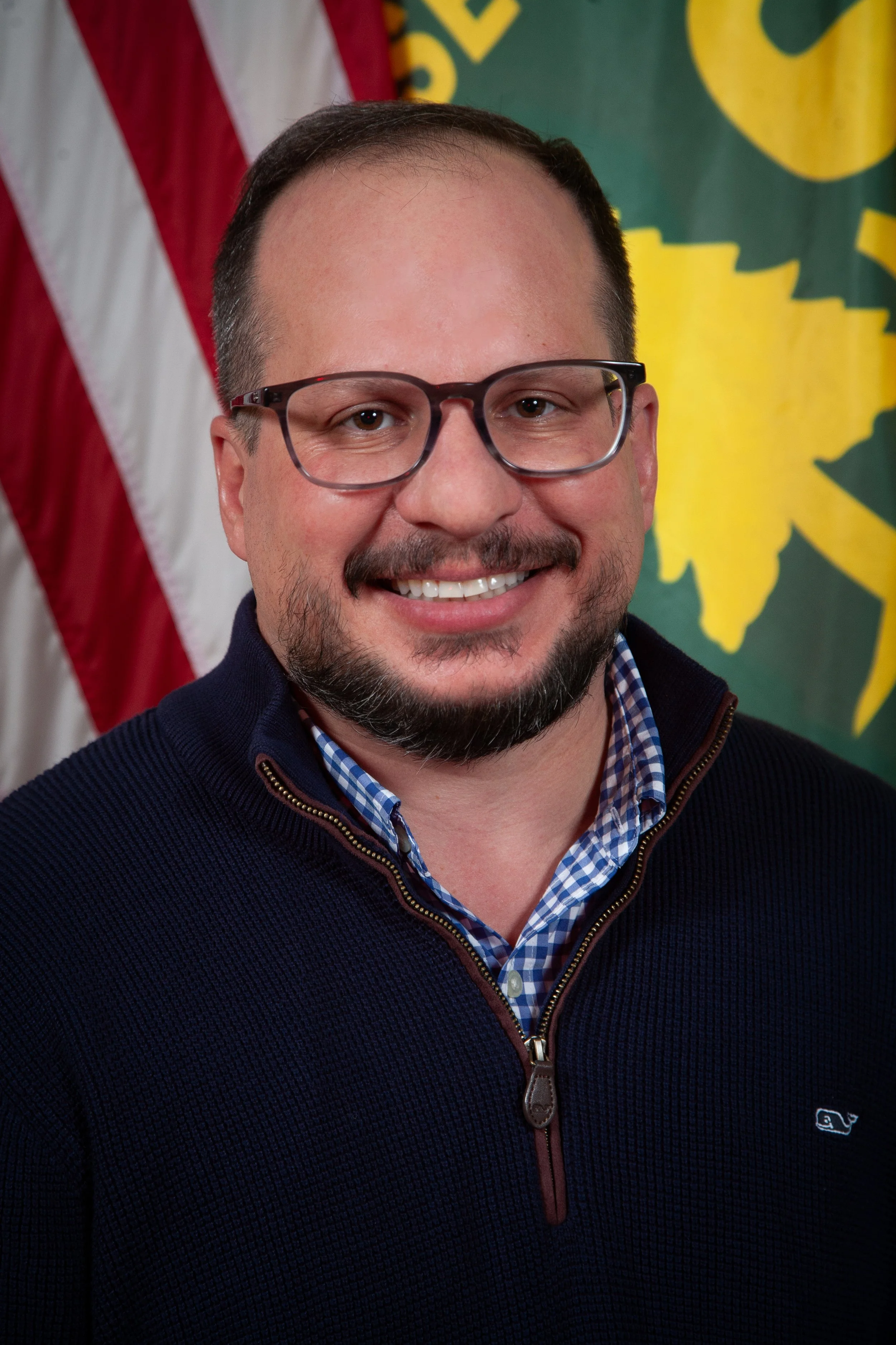 Close-up of smiling man with glasses, beard, and mustache, wearing a zipped navy sweater over a checkered shirt, background features American flag and a green banner with yellow design.