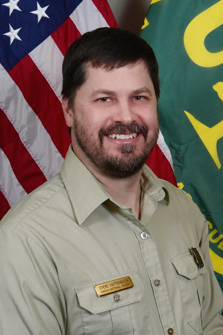 Photograph of a man with dark hair, beard, and mustache, wearing a beige uniform shirt with a name tag and badge, standing in front of American and Washington state flags.