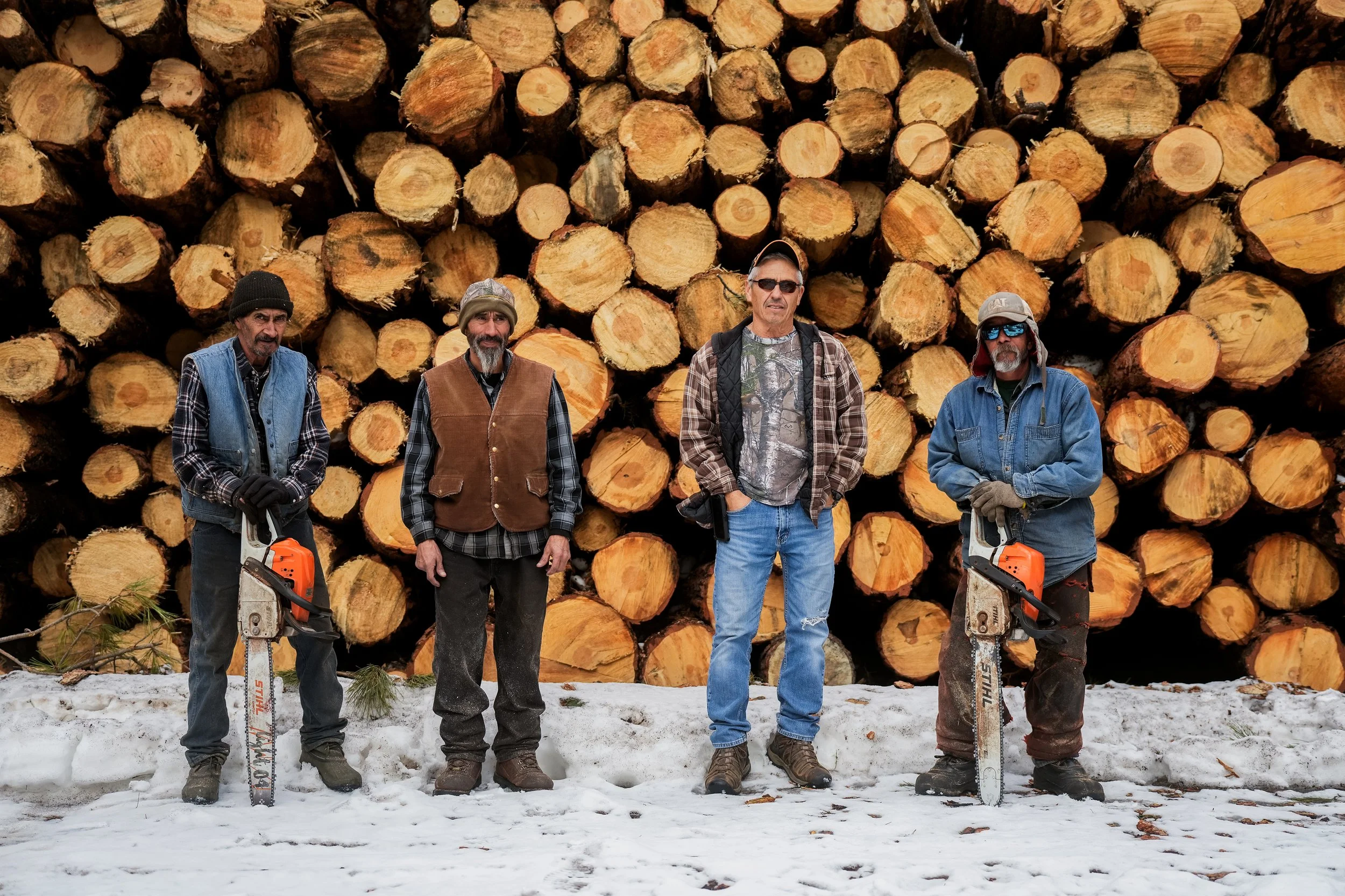 Four men in winter clothing standing on snow in front of a large stack of cut logs, some holding chainsaws.