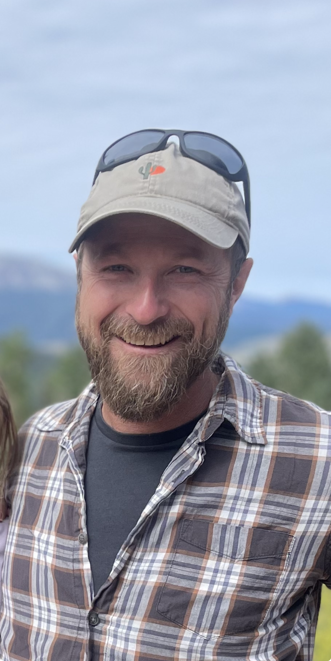 Close-up of a smiling man with a beard and light blue eyes, wearing a beige cap with a cactus embroidered on it, sunglasses on top, and a plaid shirt over a dark t-shirt, outdoors with mountains and sky in the background.