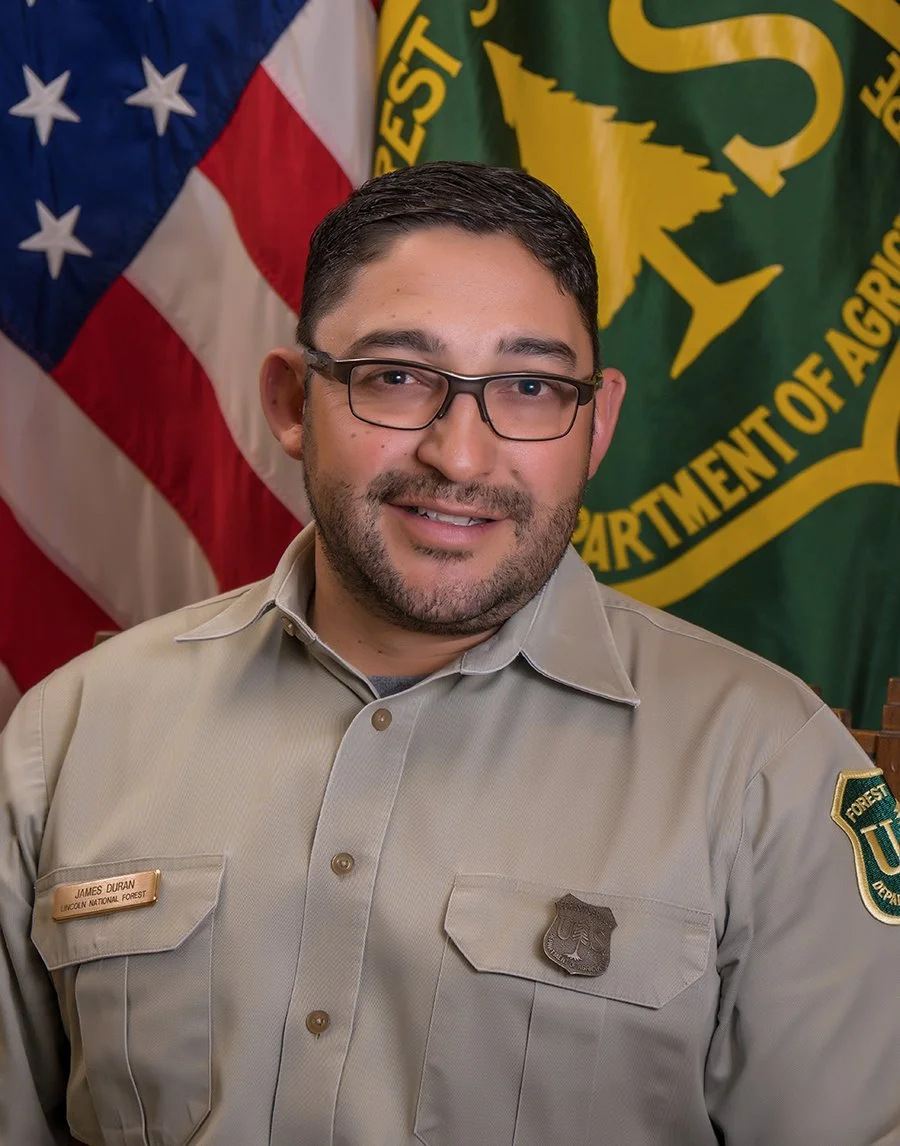A man in a forest ranger uniform standing in front of American and green vegetation department flags.