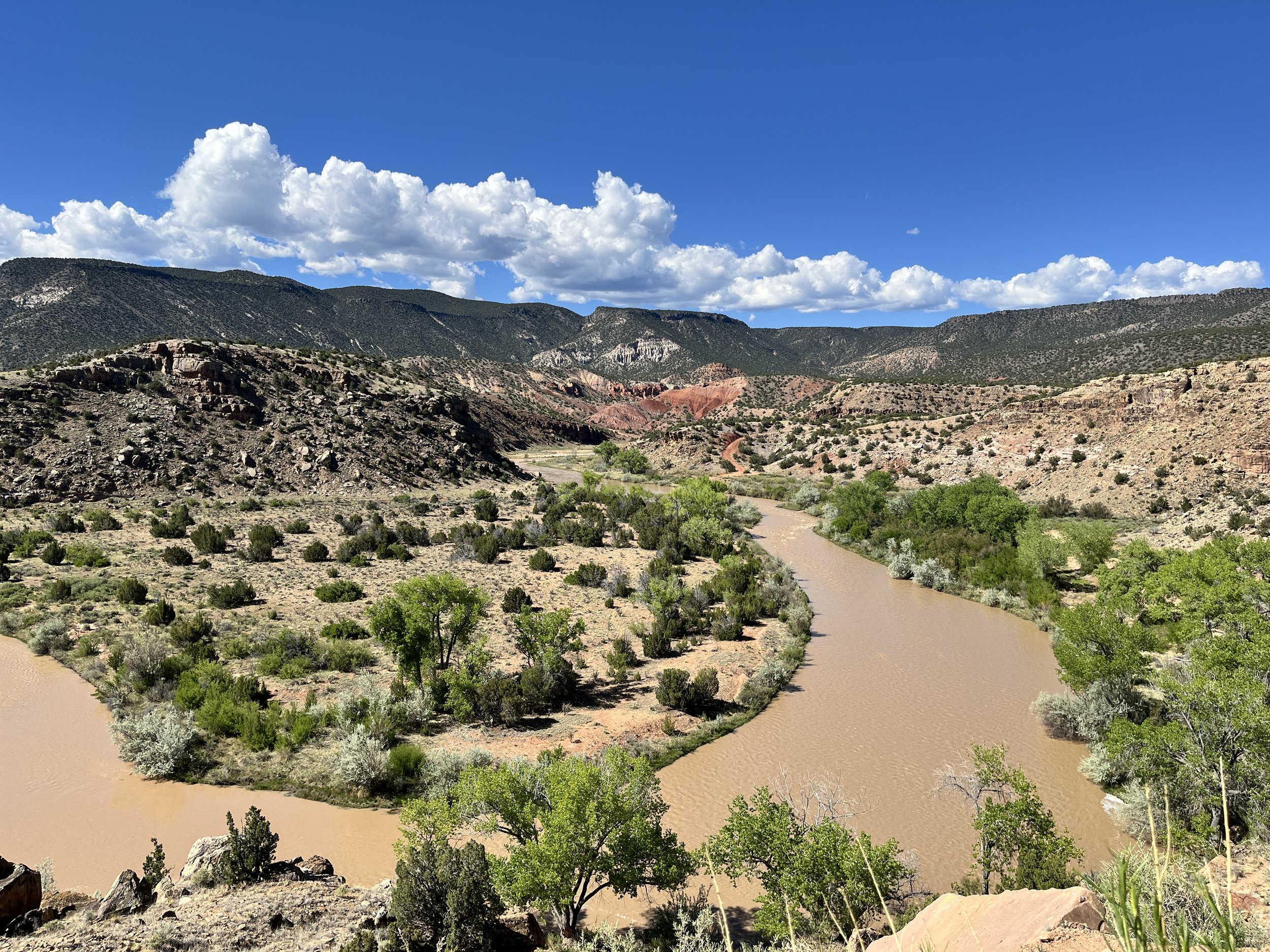A river winding through a desert landscape with green bushes and trees, surrounded by mountain ranges under a partly cloudy blue sky.