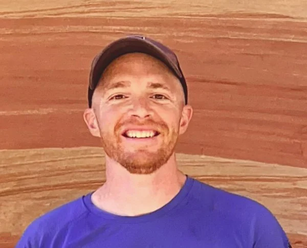 A smiling man wearing a dark cap and a blue shirt, with a wooden background.