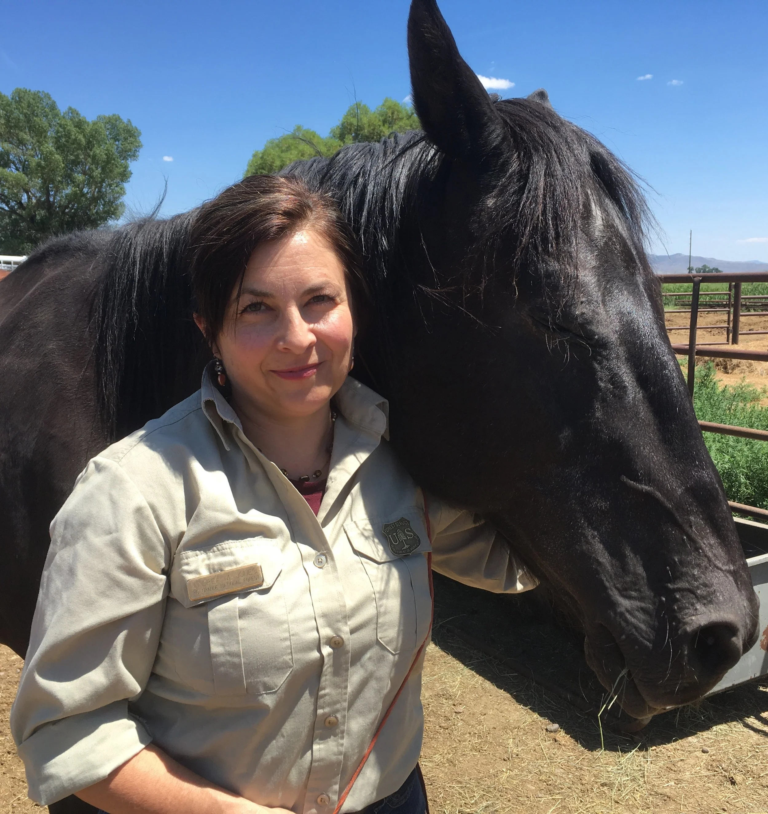 A woman with light brown hair and a beige uniform standing outdoors beside a black horse, with green trees and mountains in the background under a clear blue sky.