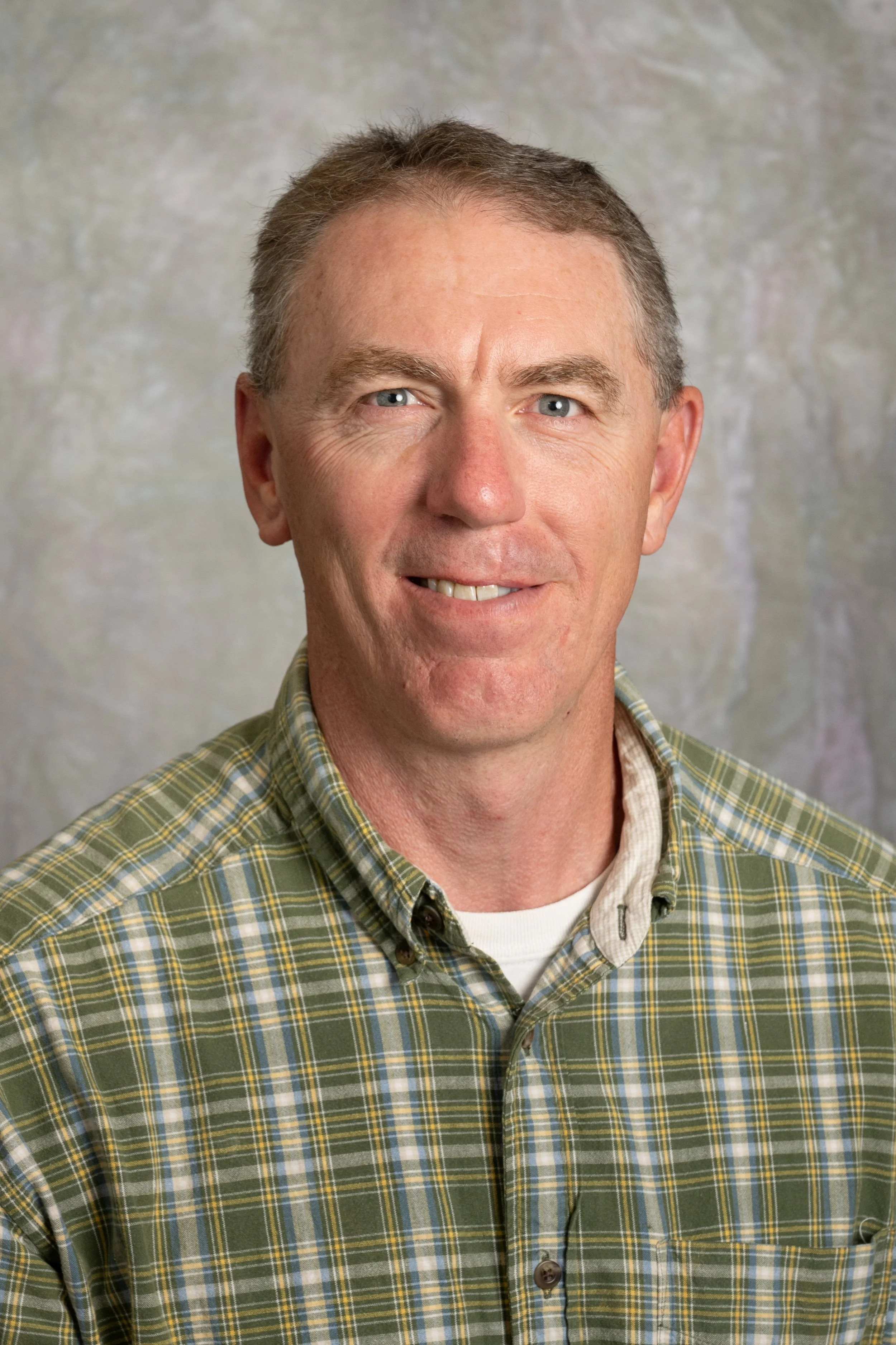 A middle-aged man with short brown hair and blue eyes, smiling slightly, wearing a green and yellow plaid shirt with a white undershirt, standing in front of a neutral textured background.