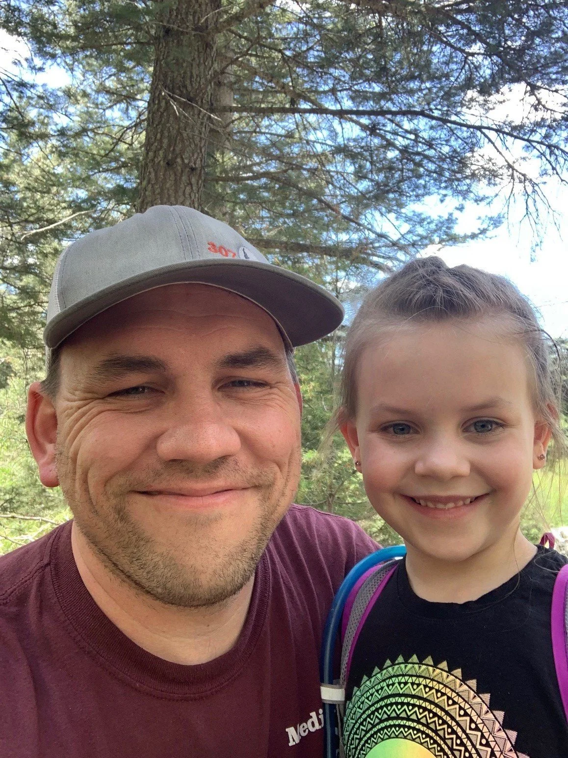 A man and young girl smiling outdoors in a forested area, with trees and blue sky in the background.