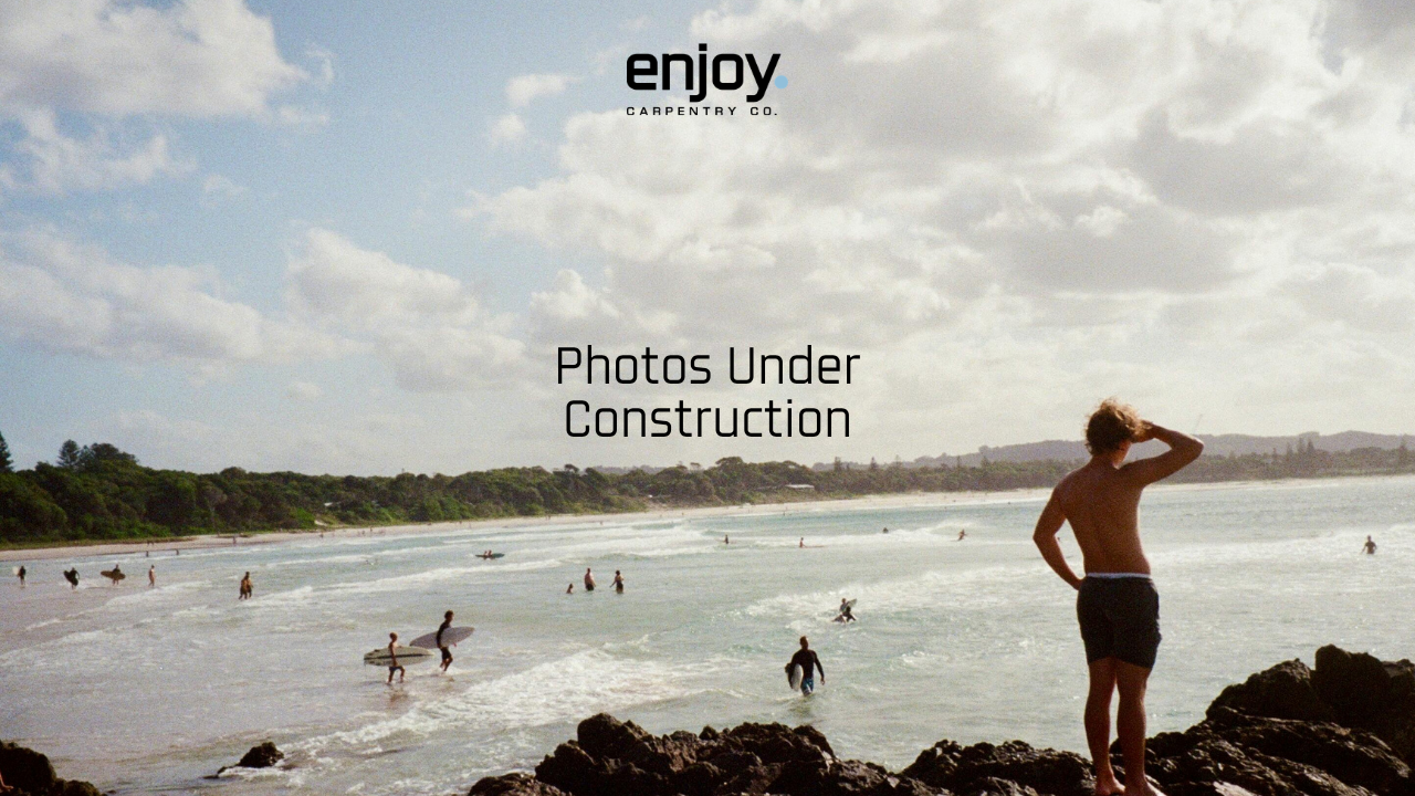 A beach scene with people surfing and swimming under a partly cloudy sky. A person stands on rocks in the foreground, overlooking the water.