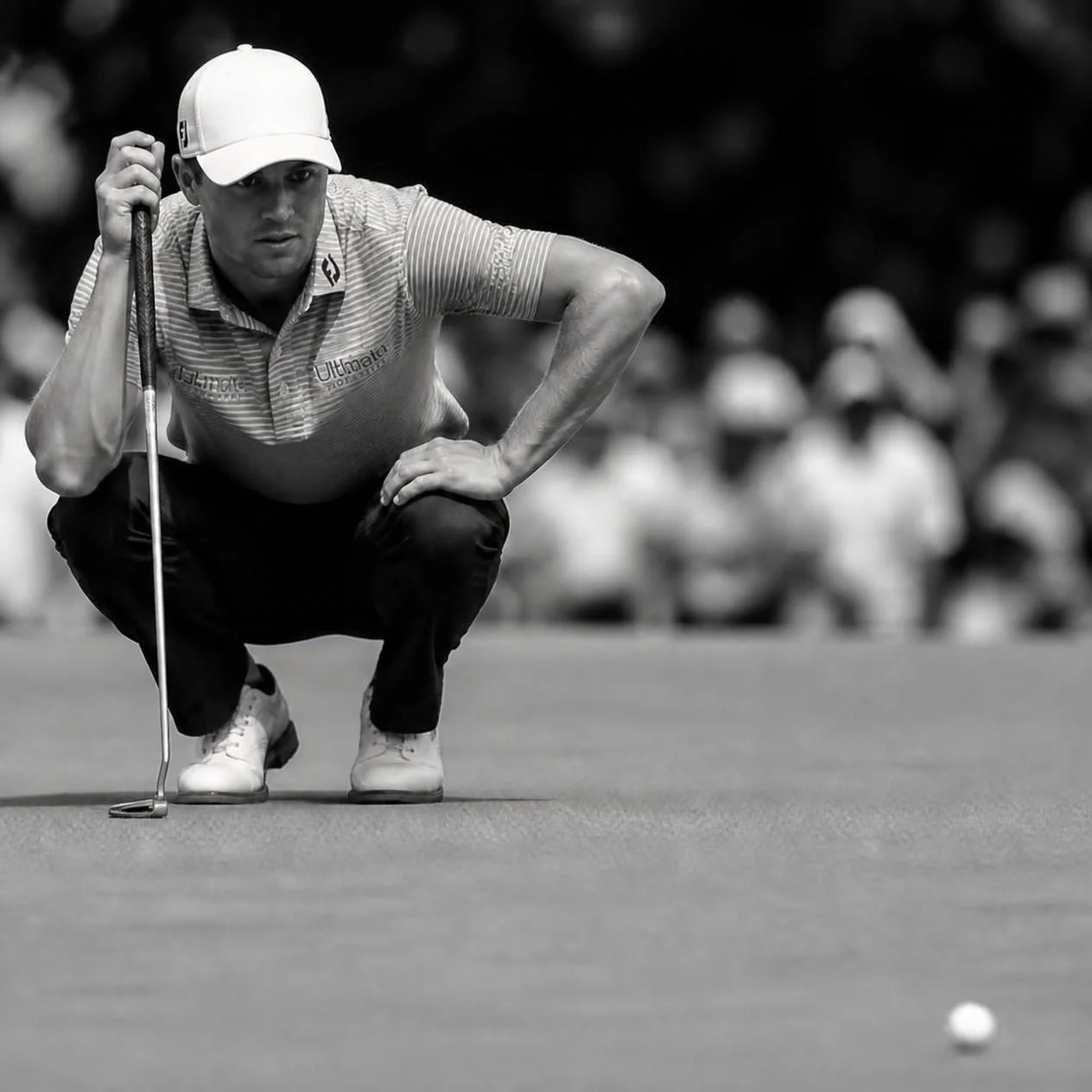 A male golfer crouching on a golf course, preparing to line up a putt with a golf ball nearby, black and white image, with spectators blurred in the background.