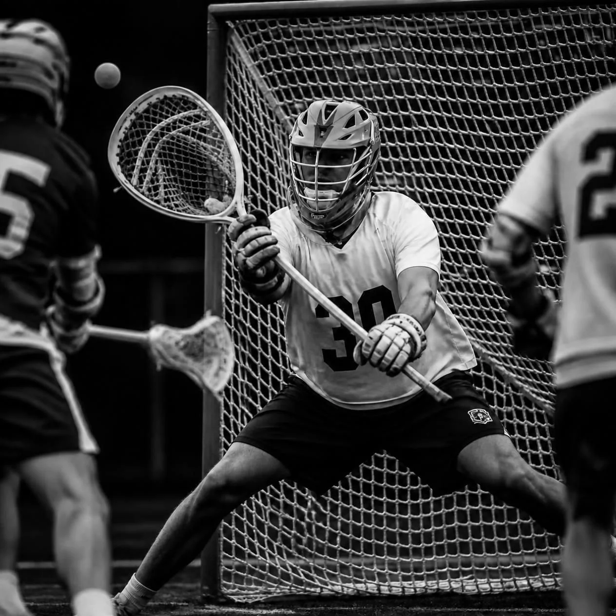 Lacrosse players engaged in a game with a goalie in a cage, preparing to defend the goal in an indoor arena.