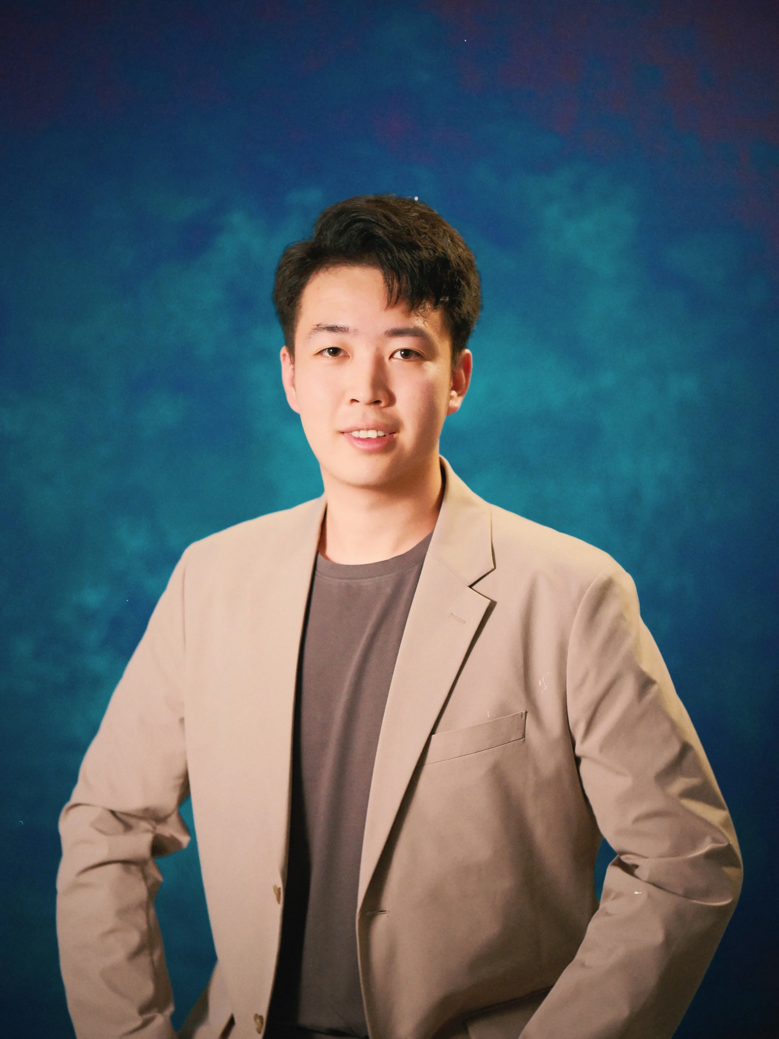 Portrait of a young man with dark hair, wearing a beige blazer over a gray shirt, standing against a blue textured background.