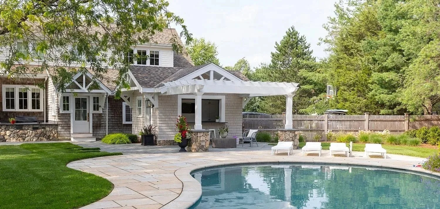 A backyard with a swimming pool, a stone patio, a house with a covered porch, surrounded by trees and a wooden fence, with lounge chairs and outdoor furniture.