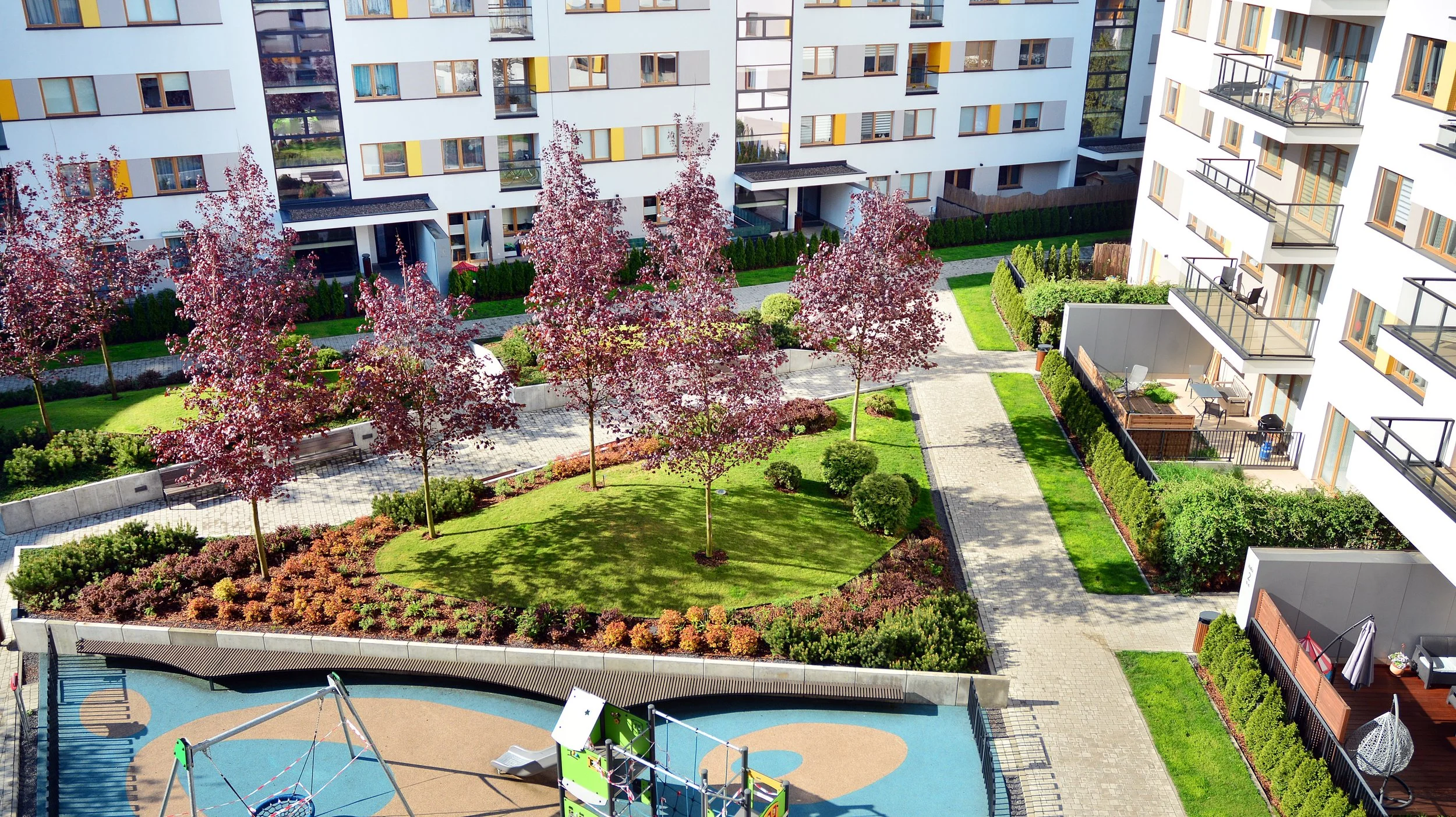 An aerial view of a modern apartment complex with a central courtyard featuring pink flowering trees and neatly landscaped green areas. The buildings have white facades with yellow and gray accent panels, balconies, and large windows. There are walkways, a small playground, and outdoor furniture visible in the courtyard.