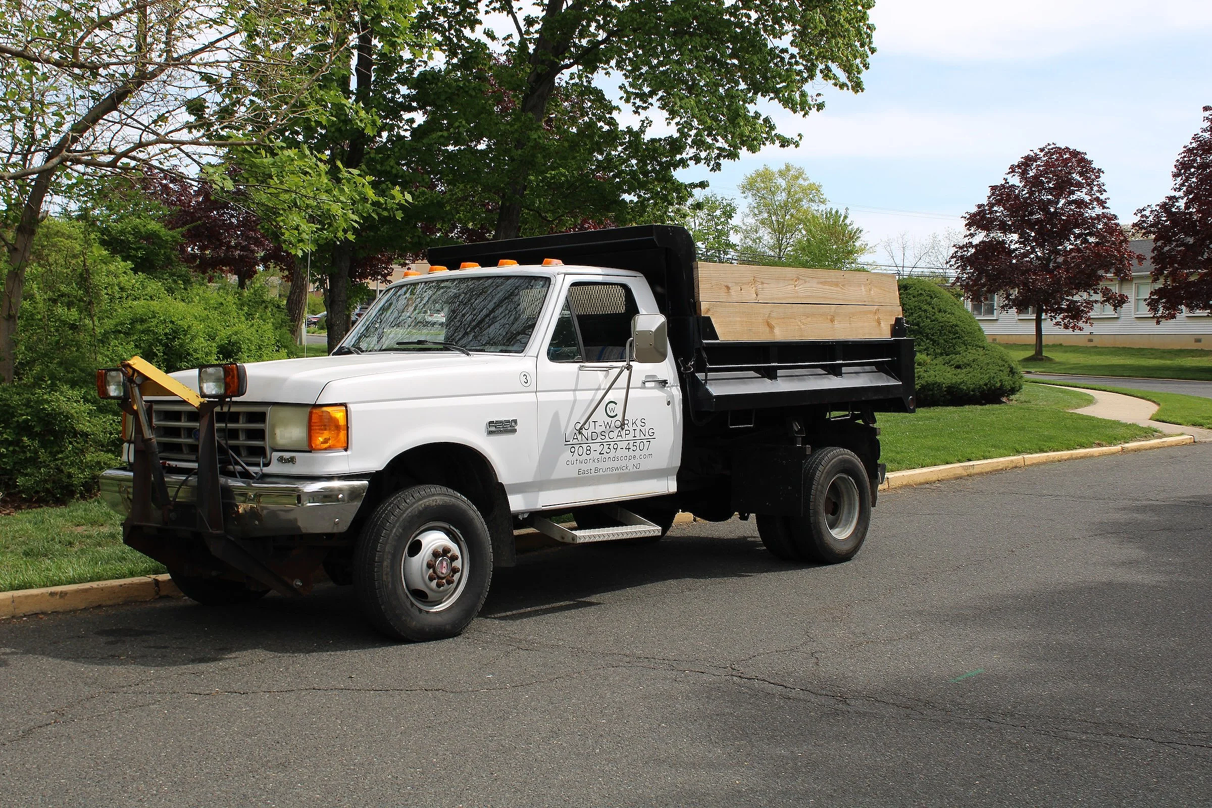 A white pickup truck with a flatbed carrying wooden planks parked on a residential street with green trees and houses in the background.