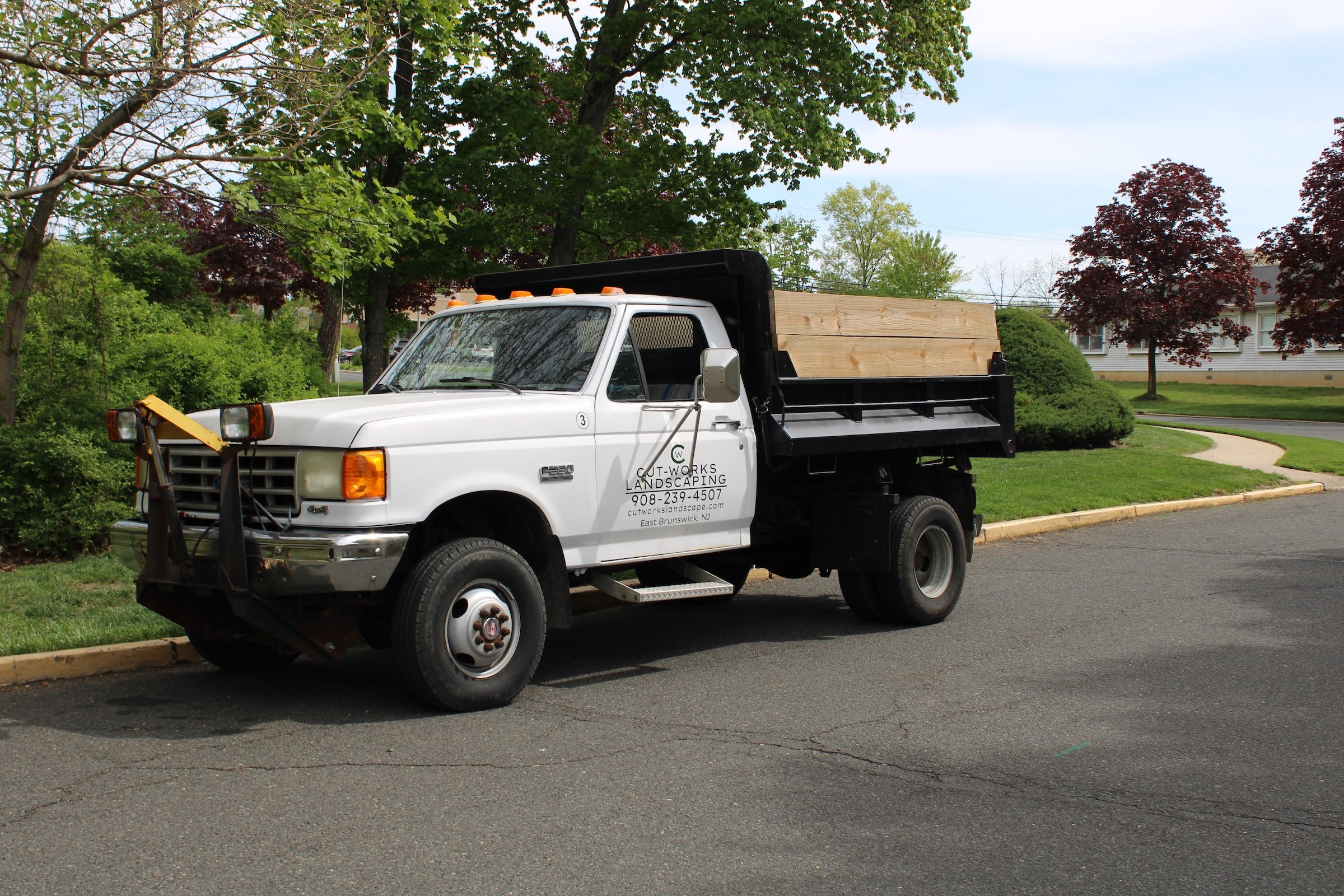 A white pickup truck with signage for a landscaping company parked on a residential street, surrounded by trees and green landscaping.