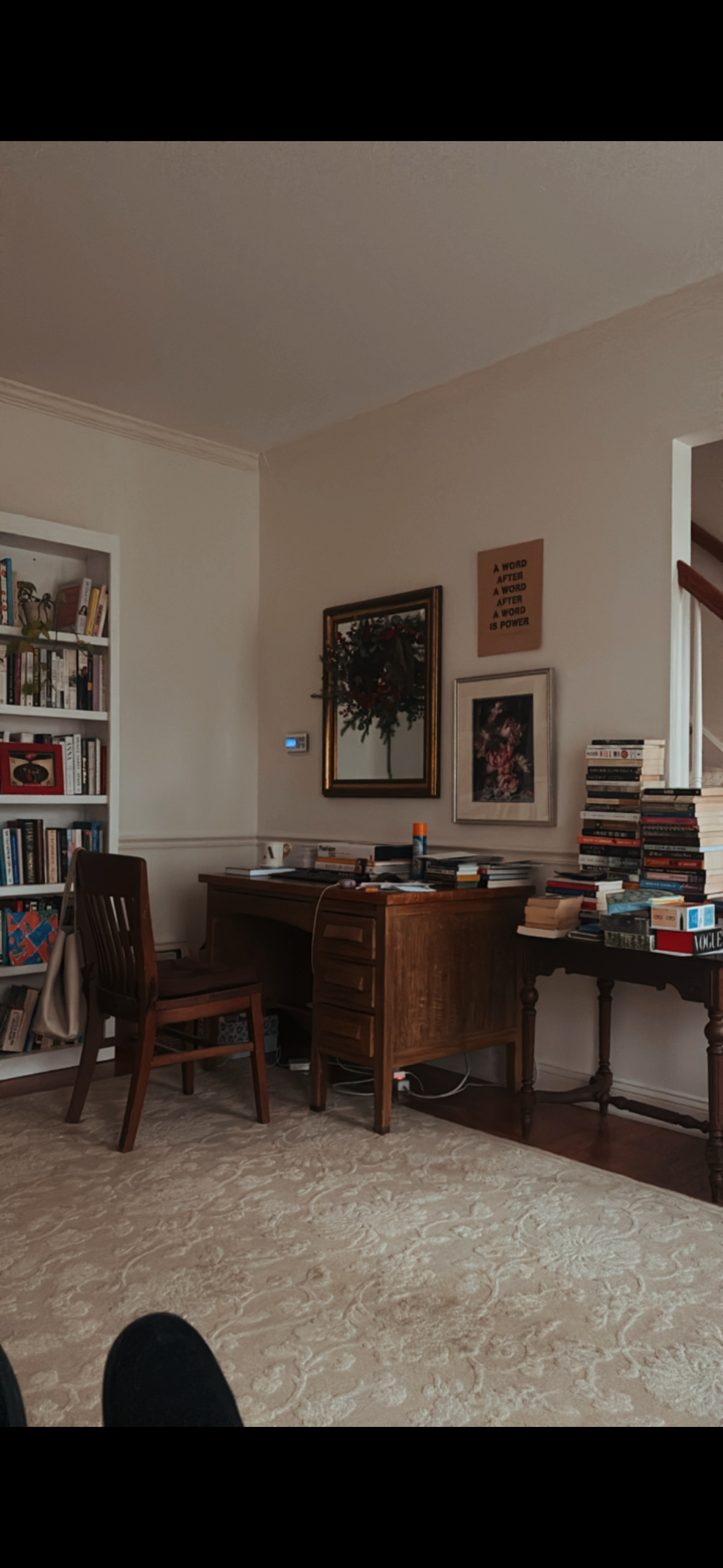 Living room with a wooden desk, bookshelf, stacks of books, pictures on the wall, and a beige patterned carpet.
