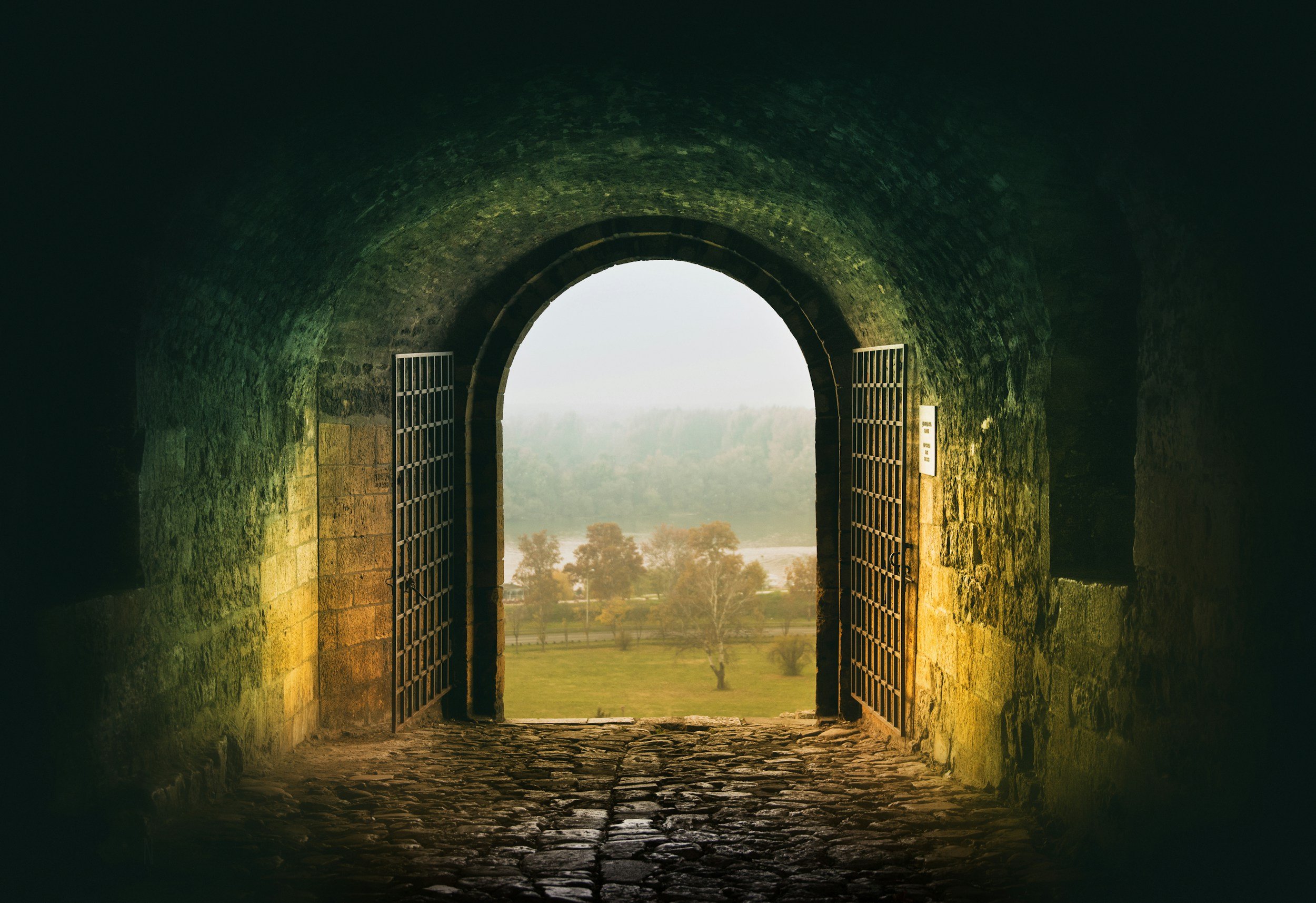 Stone archway with open gates overlooking a green landscape with trees and fields, viewed from inside a dark stone tunnel.