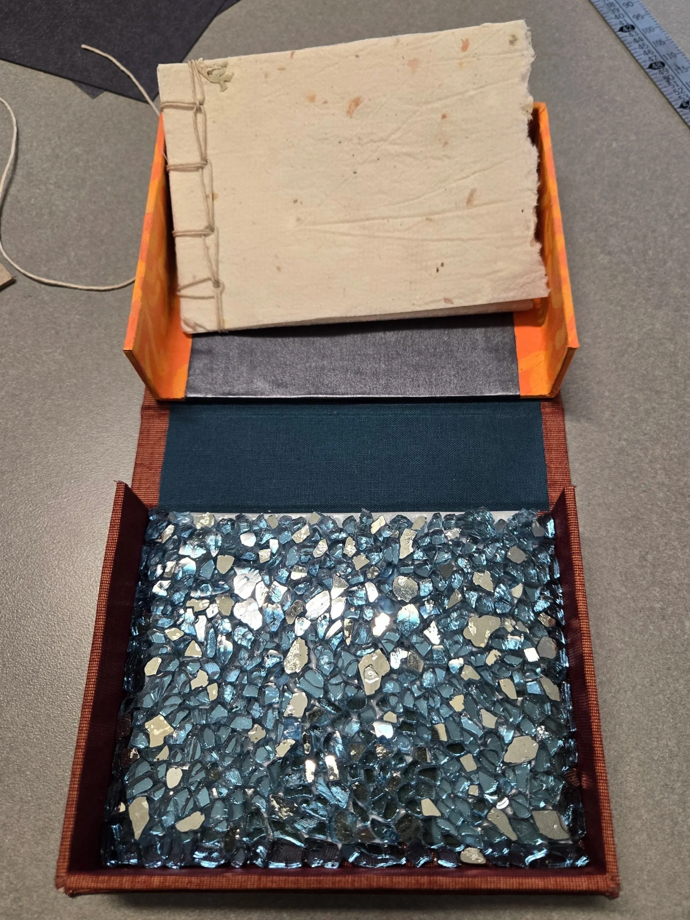 A clamshell box splayed open, revealing a stab-stitch bound booklet and glimmering blue mirror-glass