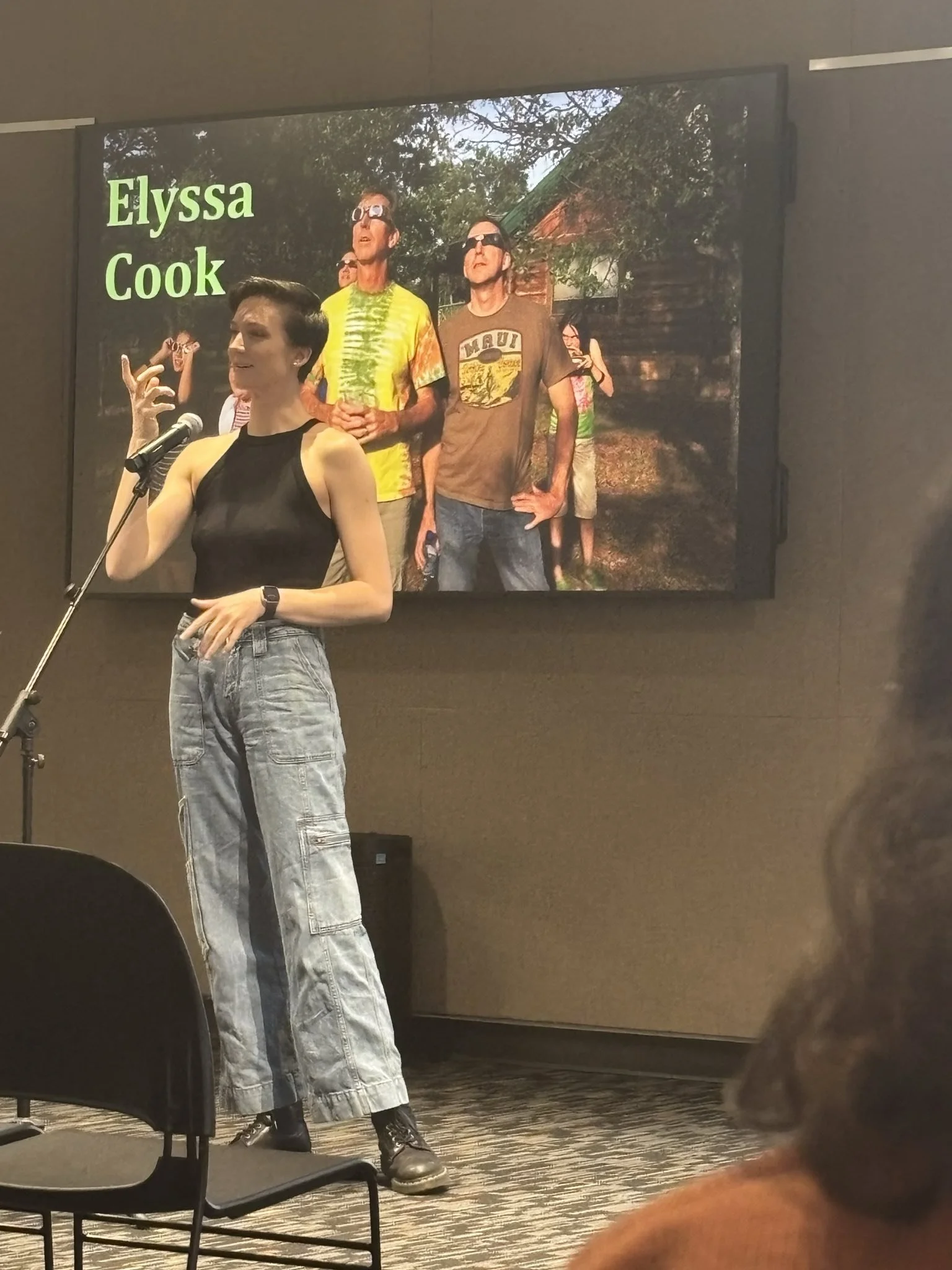 Elyssa in a black shirt standing at a microphone