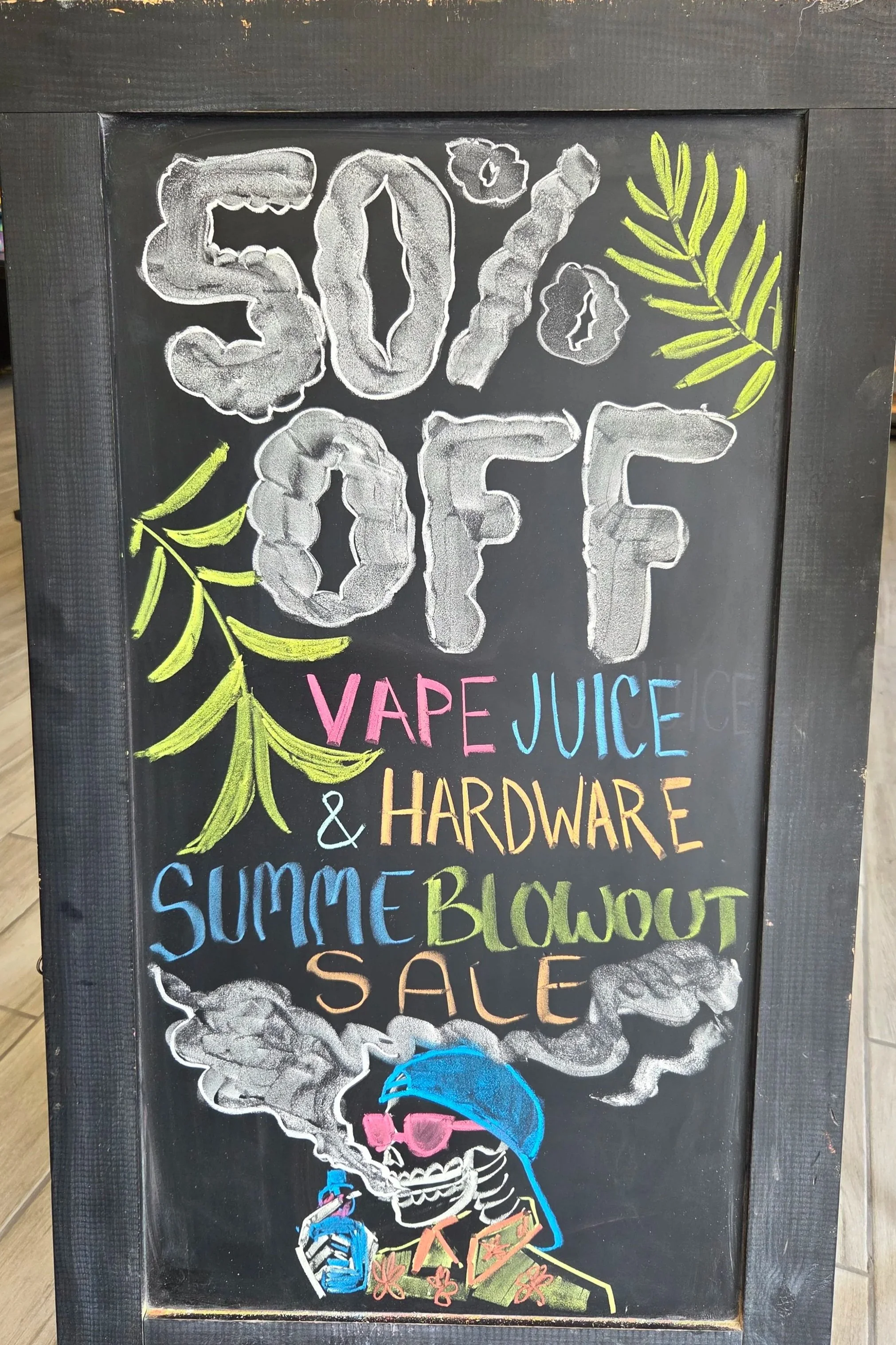 A black chalkboard sign with colorful chalk drawings and text advertising a summer sale. The top has large, white chalk letters spelling '50% OFF' with green leaves and a cloud of smoke. Below, words in various colors read 'VAPE JUICE & HARDWARE' and other sale-related phrases. The bottom features a skeleton smoking a pipe, with smoke forming additional text, and colorful vaping devices and accessories.