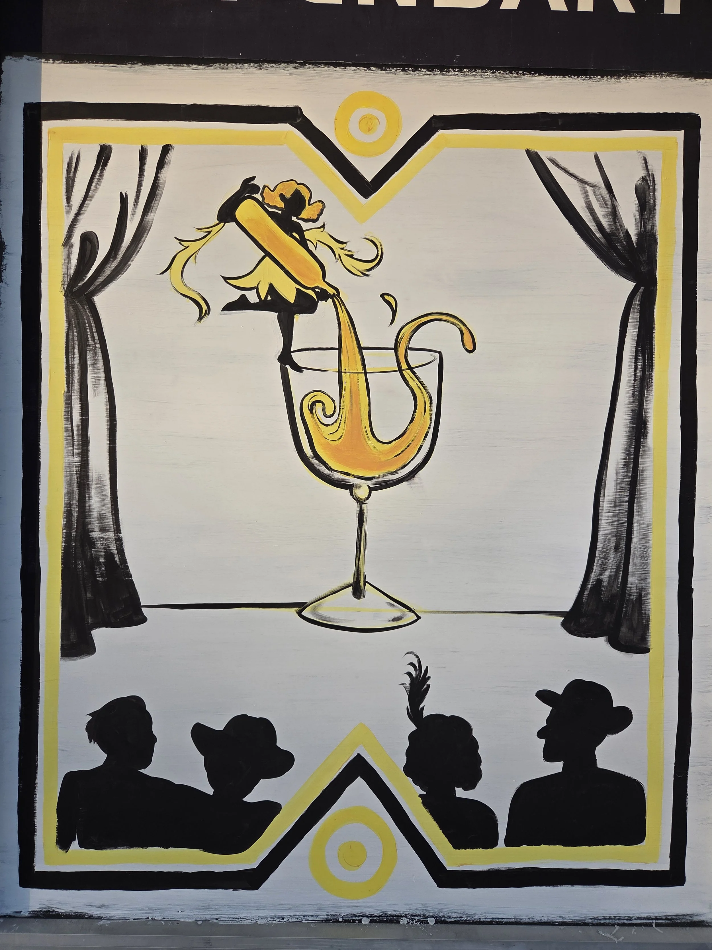A black and yellow artistic mural showing a silhouette of a woman in a dress, sitting in a large glass, with a decorative background of curtains and silhouettes of an audience in black, viewed from behind.