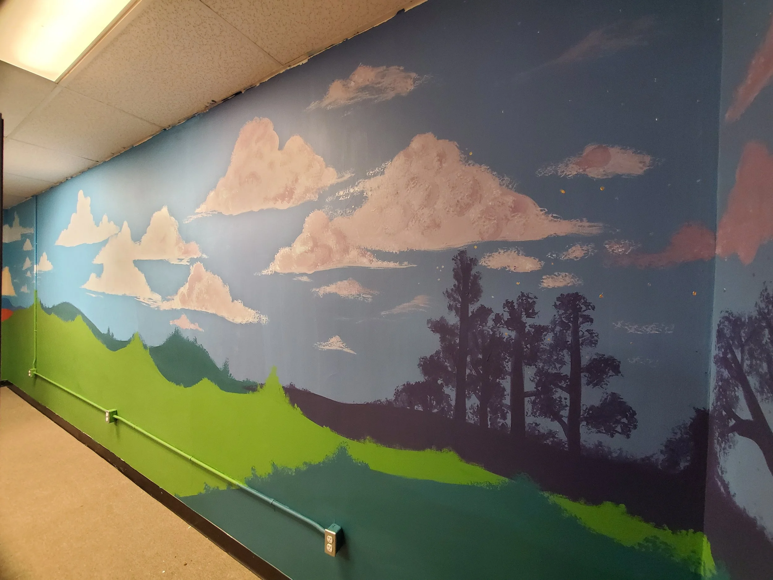 A mural painting of a landscape with green hills, trees, blue sky, and fluffy clouds on a wall in an indoor space.