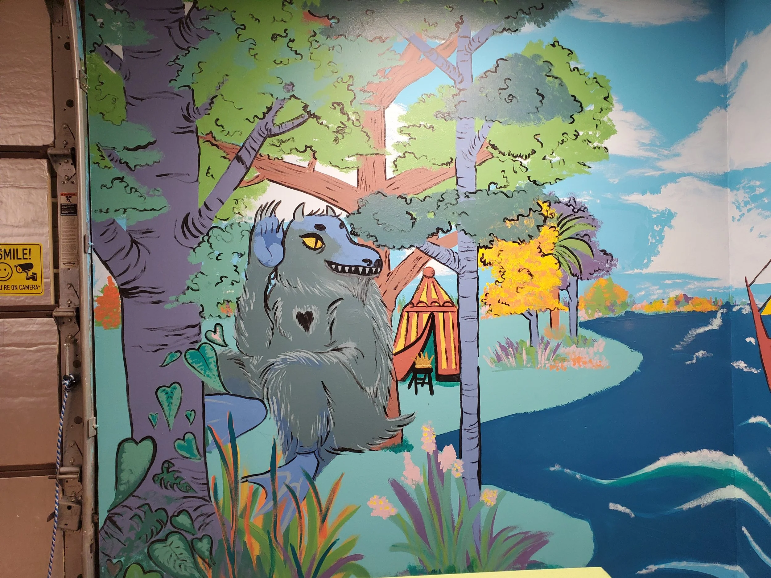 Colorful mural featuring a cartoonish goat standing among green grasses and purple plants near a body of water, with trees and a bright blue sky with white clouds in the background.