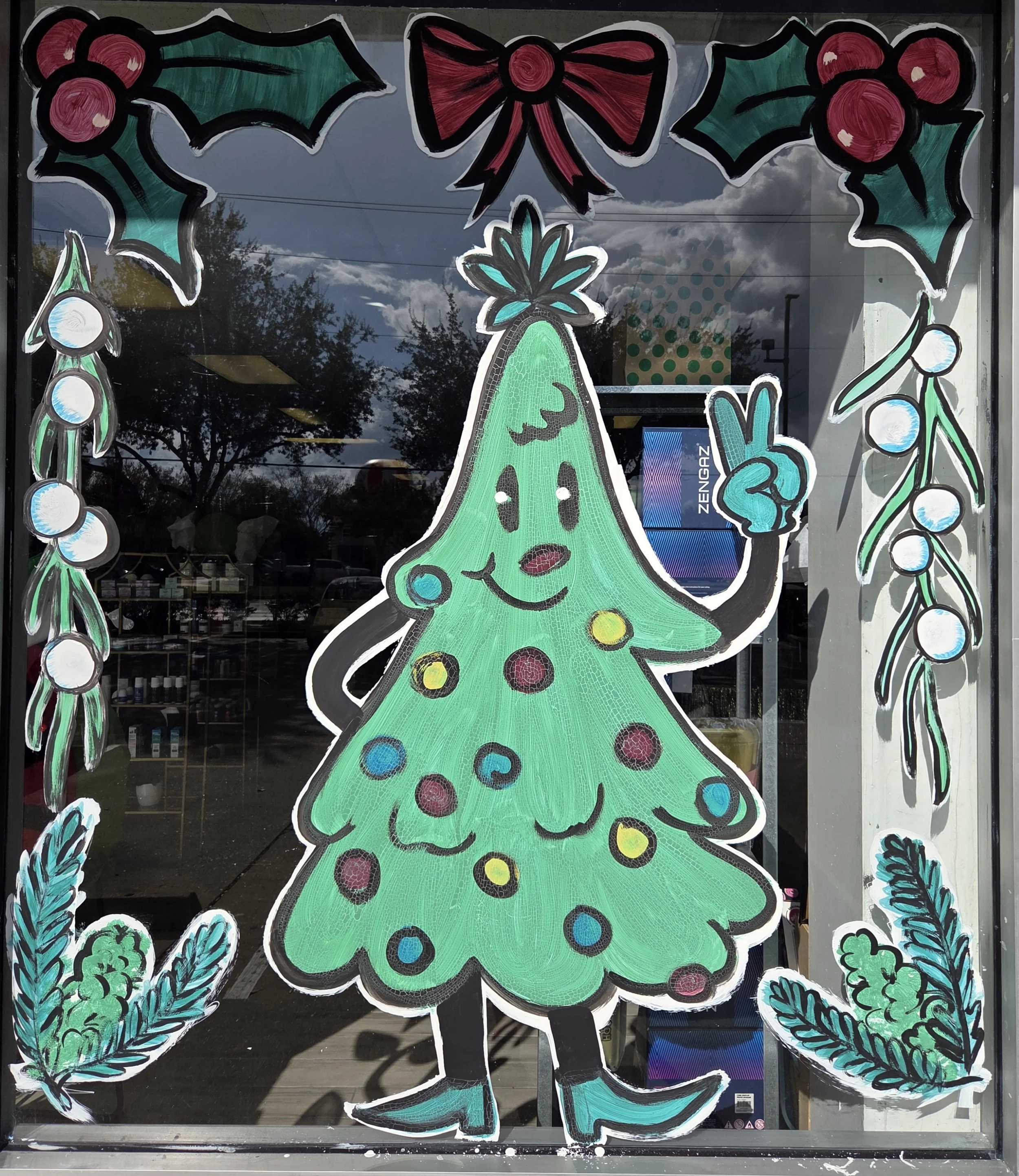 Decorative holiday window display featuring a large smiling Christmas tree with colorful ornaments, a red bow, holly, and mistletoe. The tree has a face with eyes, a nose, and a mouth. Surrounding the tree are green holly leaves with red berries and 