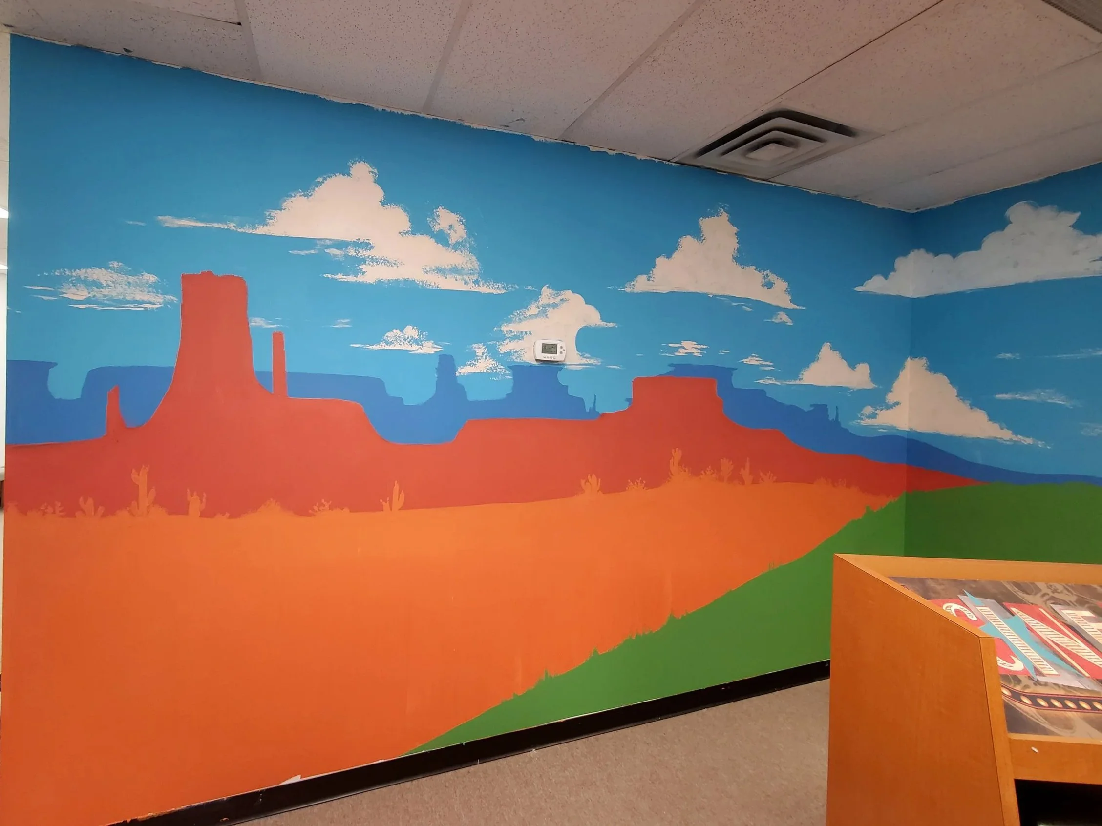A colorful mural depicting a desert landscape with orange mesas, blue sky, and white clouds, painted on a wall indoors with a drop ceiling and a thermostat on the wall.