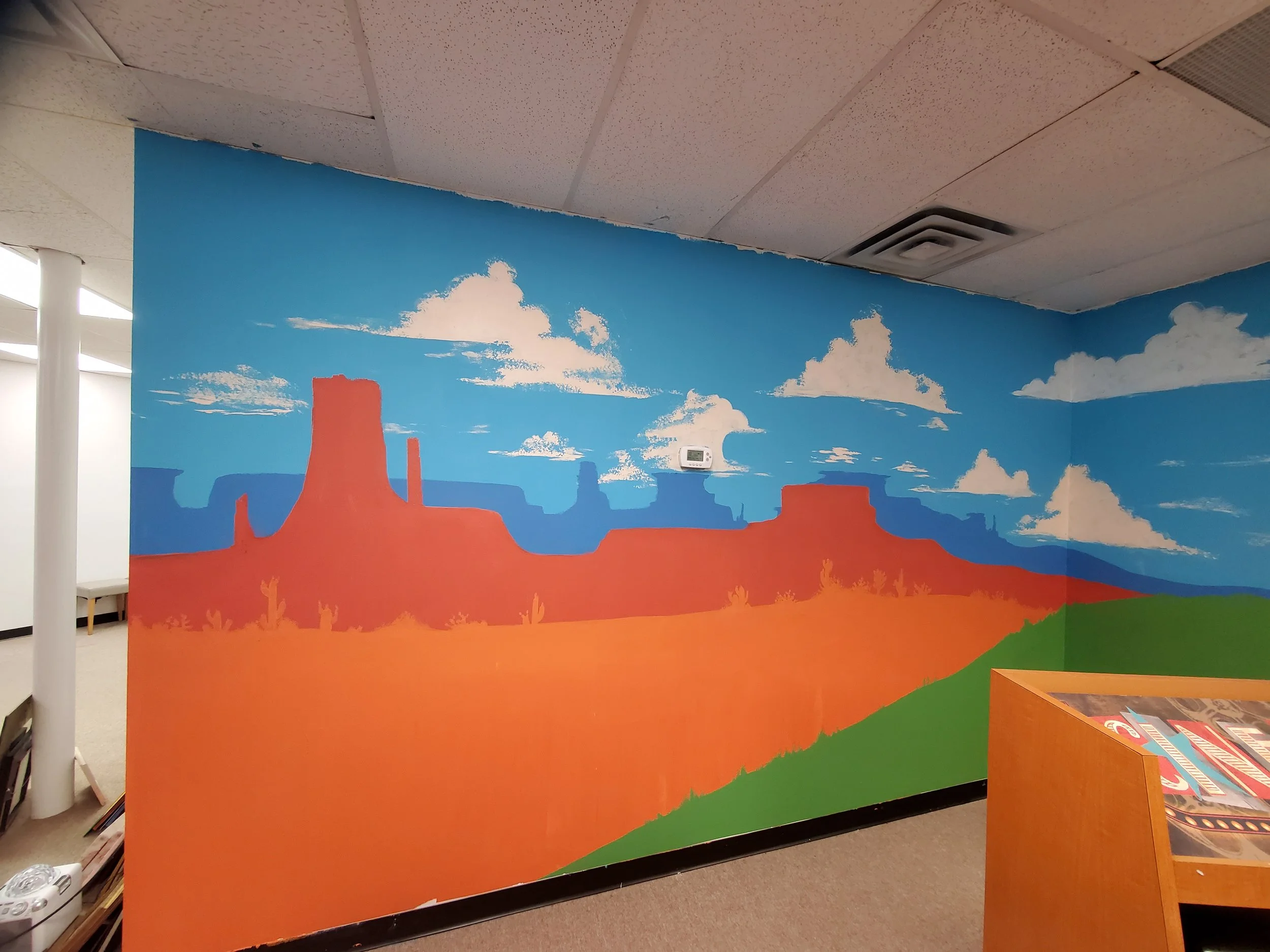 Colorful mural depicting a desert scene with blue sky, clouds, and orange rock formations, in an indoor space.