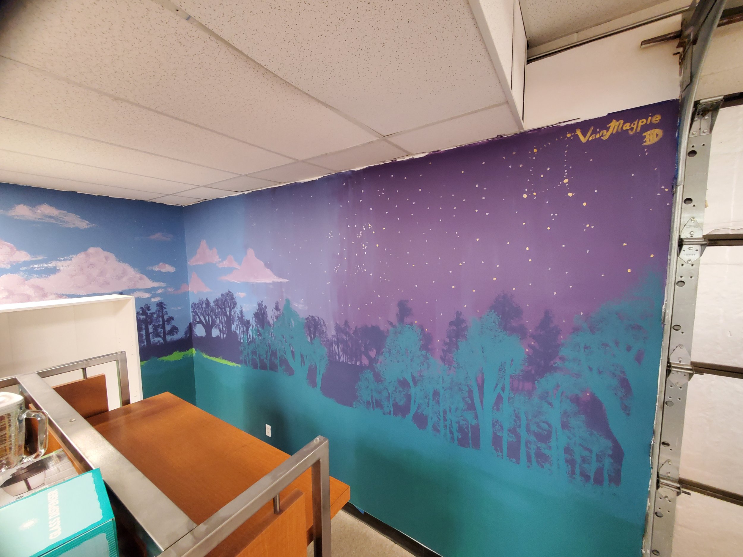 Indoor scene with a wall mural of trees and clouds, and a small section of furniture and shelves visible in the foreground.