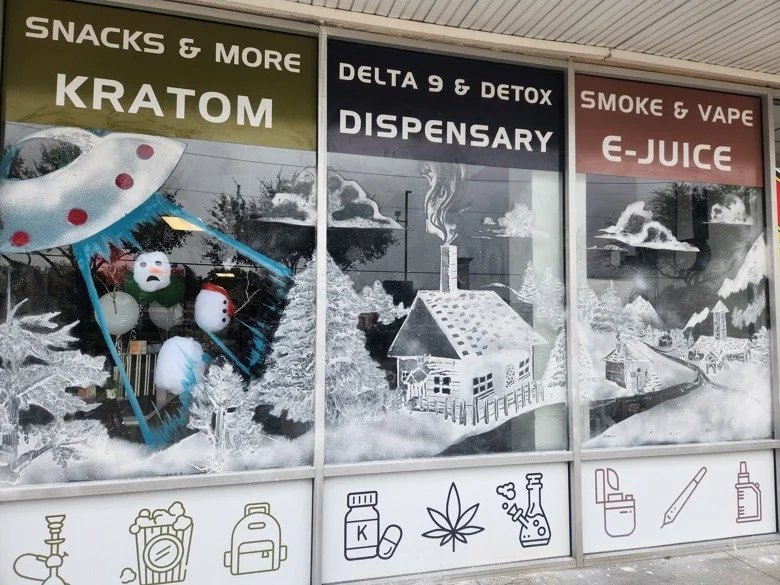 Storefront window with signs for kratom, delta 9 and detox, dispensary, smoke and vape. Window is a spray-snow painted scene of a winter countryside with houses, trees, clouds, and a small village. In the foreground a UFO is abducting a snowman