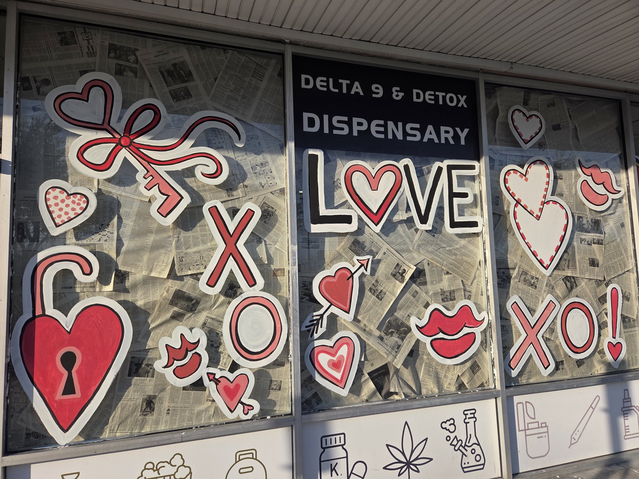 Storefront window with signs that say 'Delta 9 & Detox Dispensary' and display various red and white heart and love-themed illustrations, with newspaper clippings behind the signs.