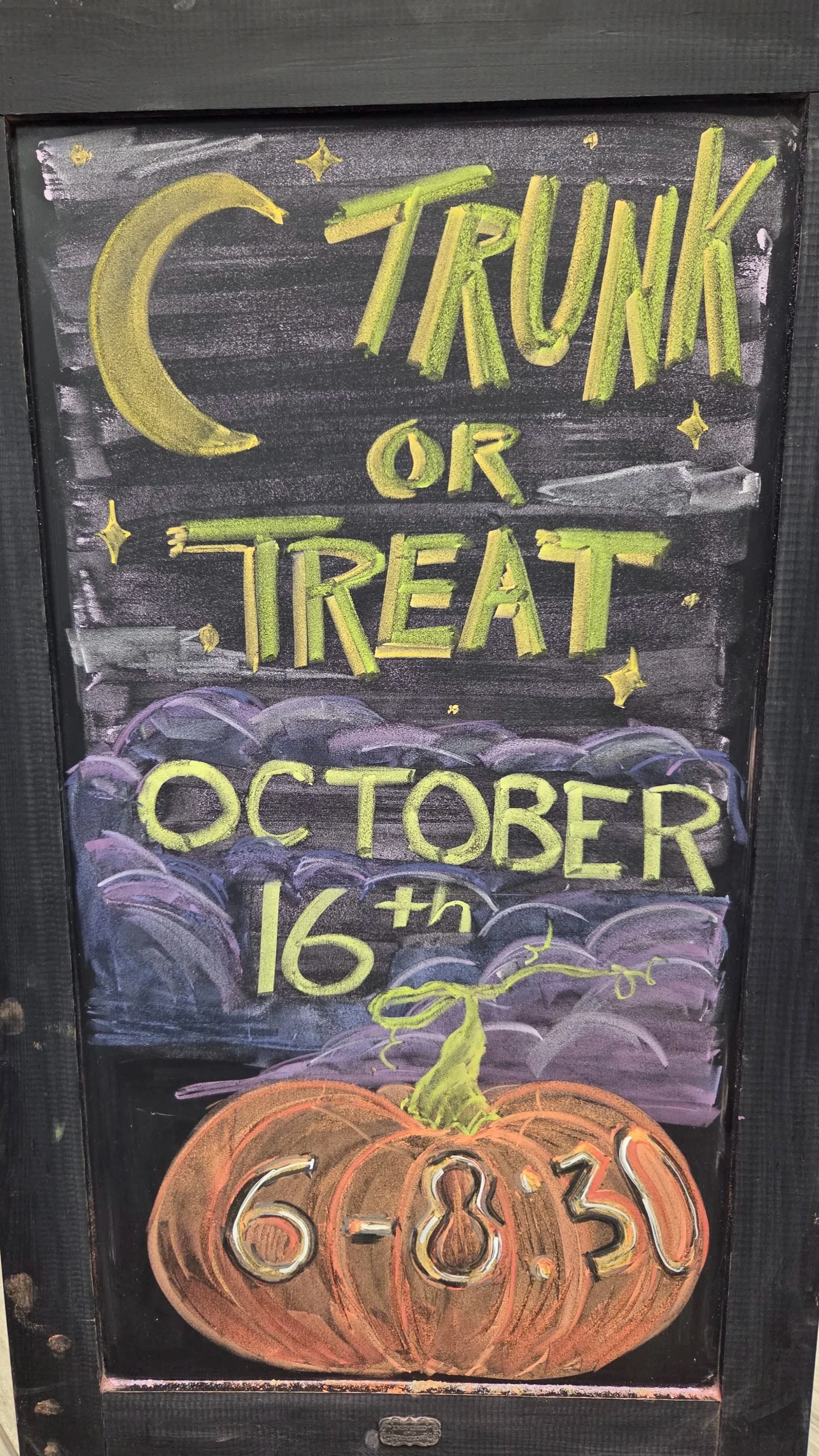 Signboard advertising a Halloween event with a moon, stars, purple clouds, a pumpkin with carved numbers, and text in yellow and green about Trunk or Treat on October 16th.