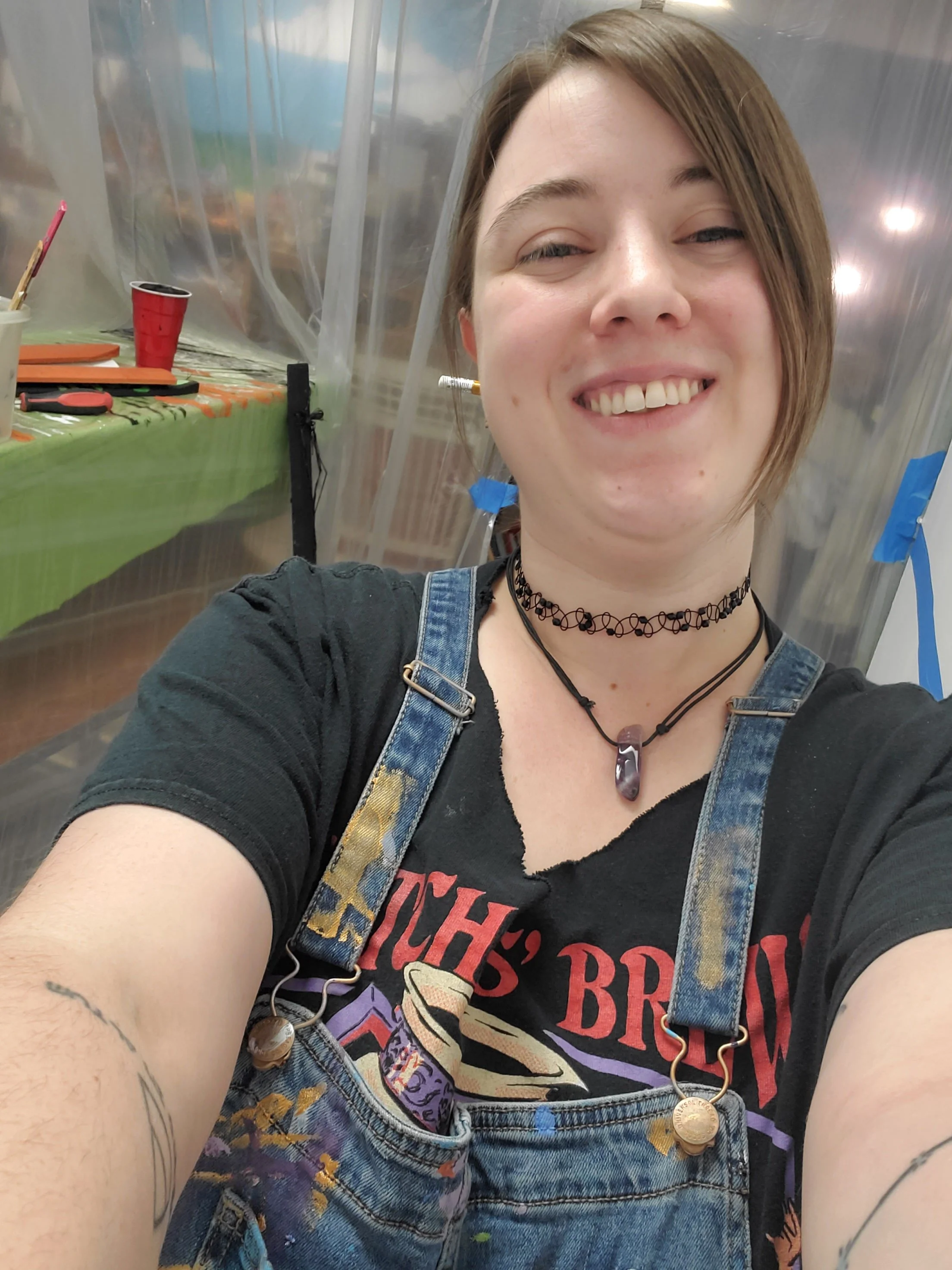 Close-up selfie of a young woman with short brown hair, smiling, wearing a black graphic t-shirt, denim overalls, a choker, and layered necklaces, captured in a space with a metallic background and a table with art supplies.