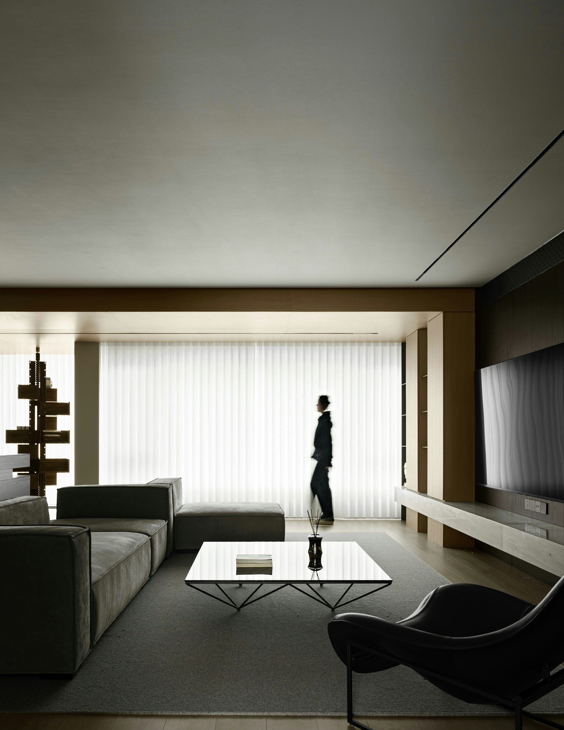 Modern living room with gray velvet sofa, black lounge chair, glass coffee table with black metal frame, gray carpet, white vertical blinds, and a large flat-screen TV mounted on a wooden wall.