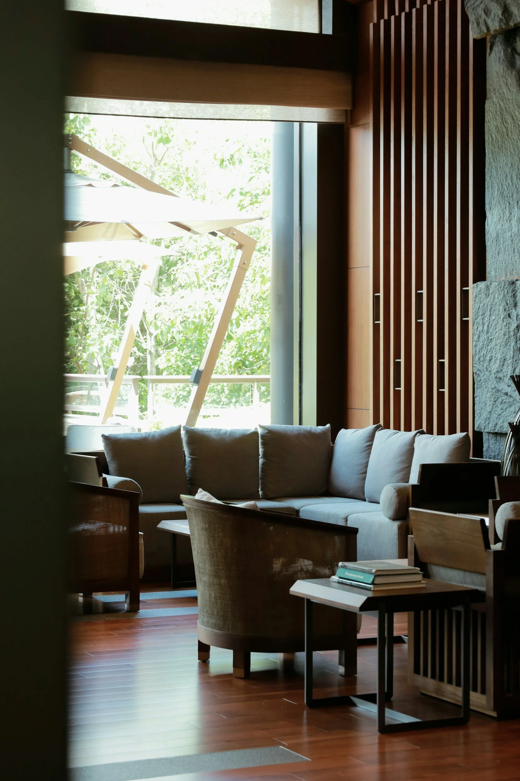 Living room with beige sectional sofa, wooden armchair, and coffee table, near large window with green trees outside.
