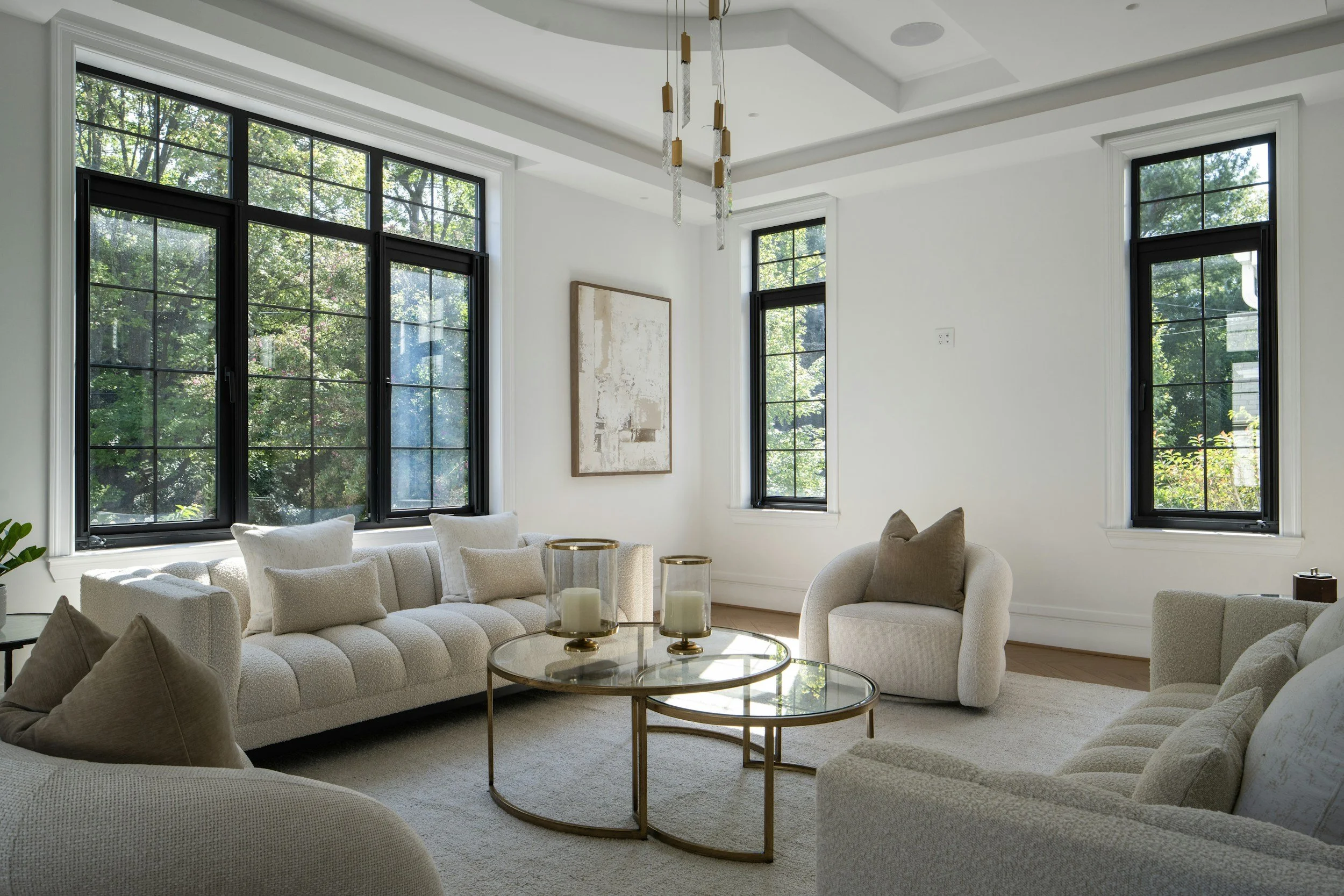 Living room with large black-framed windows, white walls, beige sectional sofa, round glass coffee table with candles, and wall art. Trees visible outside the windows.