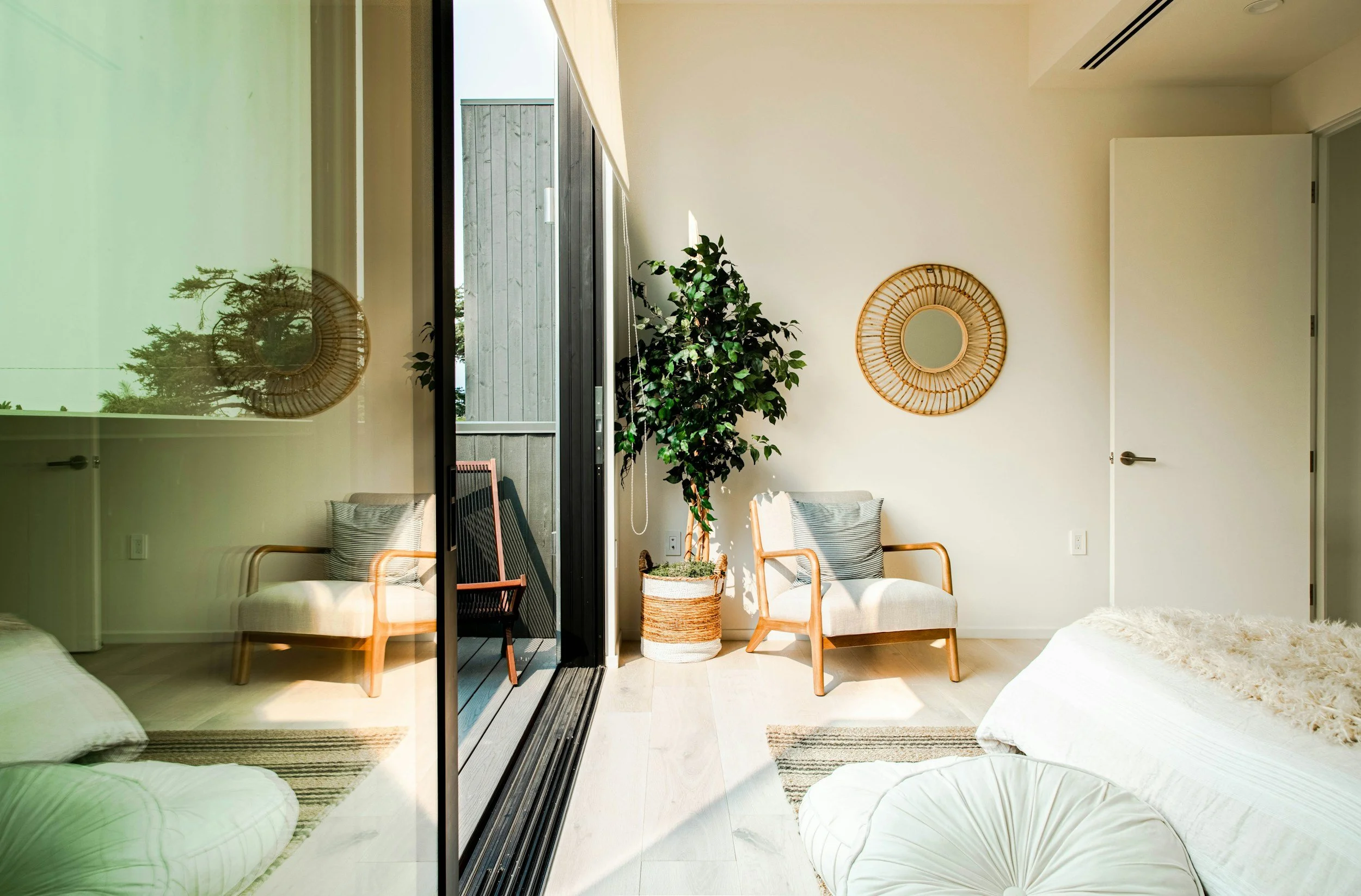 Modern living space with large glass door leading to an outdoor balcony. Inside, there are chairs with striped cushions, potted plants, and two round wall mirrors made of rattan. The room is decorated in light neutral tones with minimalistic style.