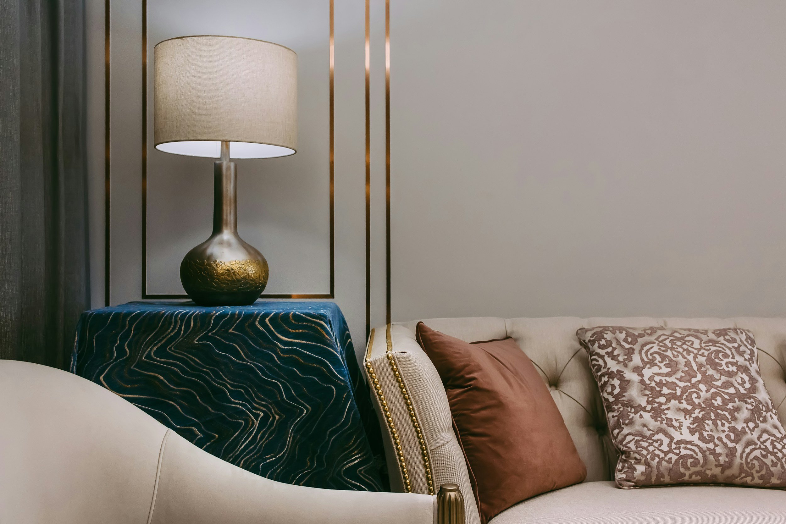 A beige sofa with patterned and solid-colored pillows, a navy blue throw with gold accents, and a gold and beige table lamp on a side table in a modern living room.