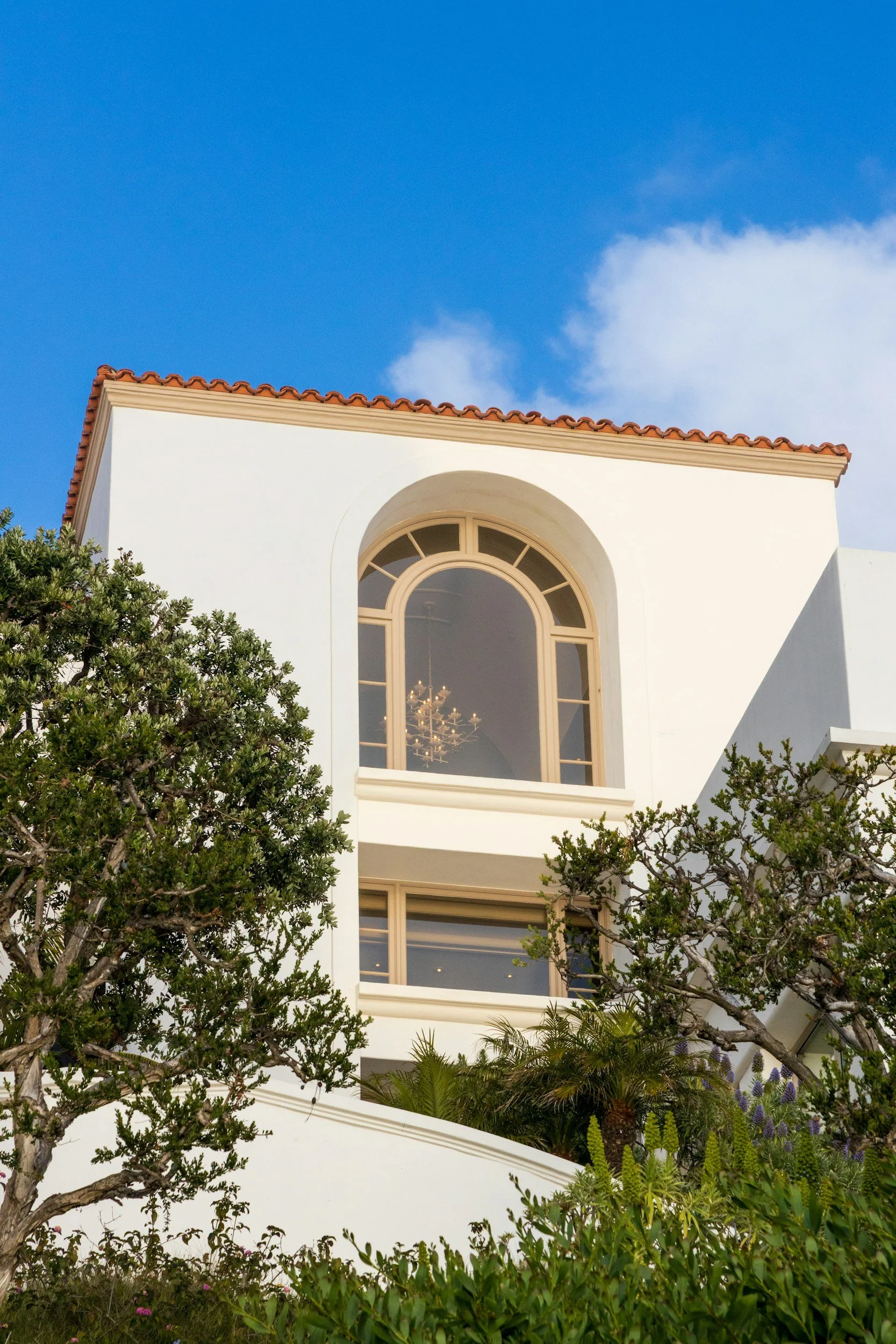 A white building with a red-tiled roof, arched window, and lush greenery in the foreground, against a bright blue sky with a few clouds.