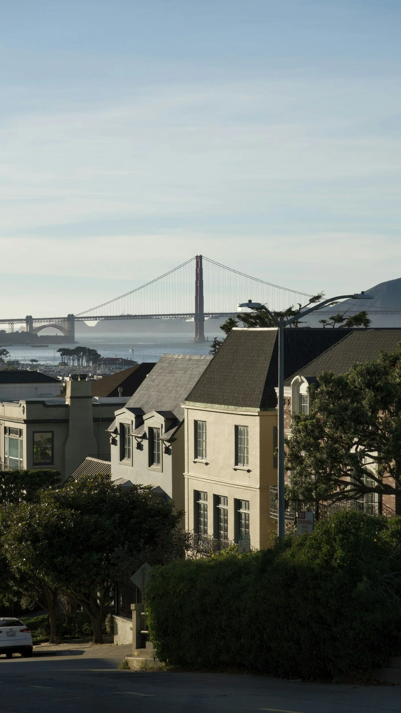 View of residential houses in the foreground with a large body of water and the Golden Gate Bridge in the background, under a partly cloudy sky.