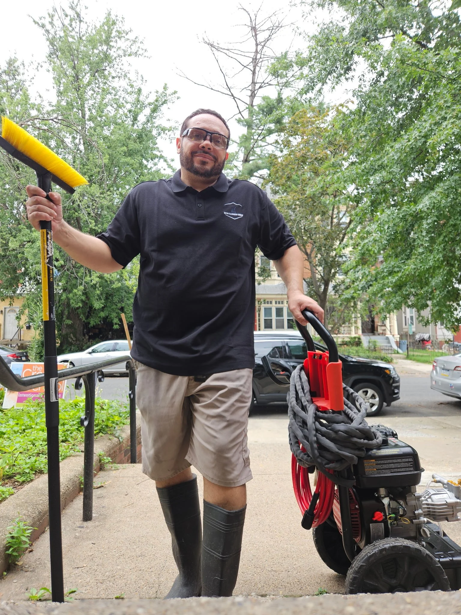 A man wearing safety glasses, black T-shirt, beige shorts, and rubber boots is standing outdoors in front of a gas-powered weed trimmer, holding the device with one hand. There are green trees, cars, and residential buildings in the background.