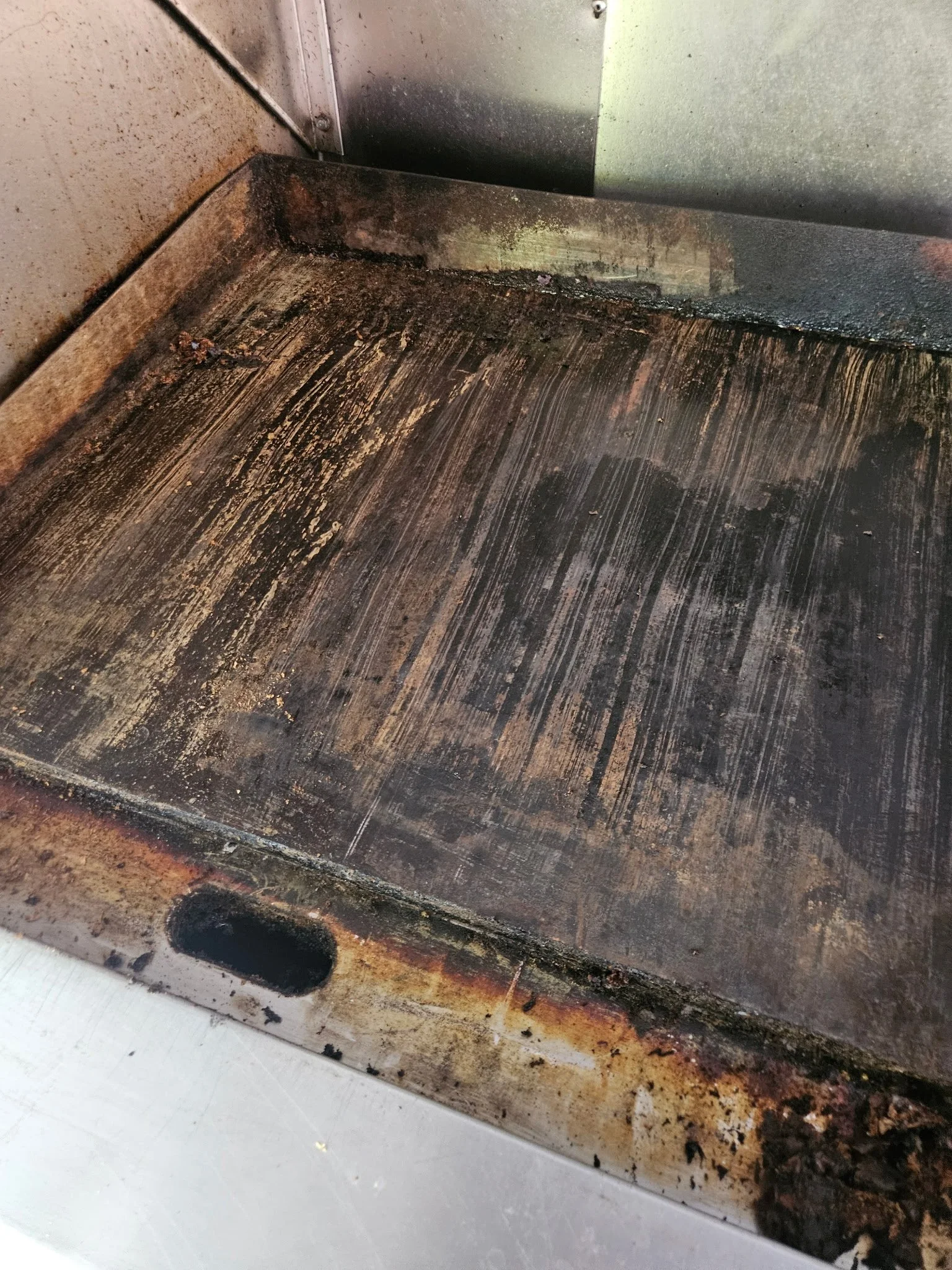 A close-up of a dirty, greasy, and burnt oven or appliance surface, with black grime and rust around the edges and at the base.