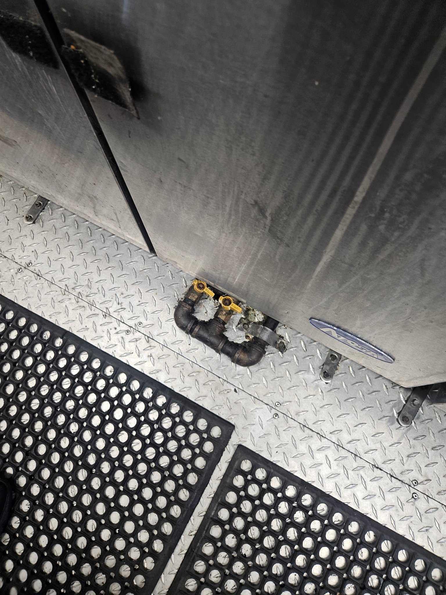 Close-up of a metallic industrial floor with black rubber mats and visible plumbing pipes.