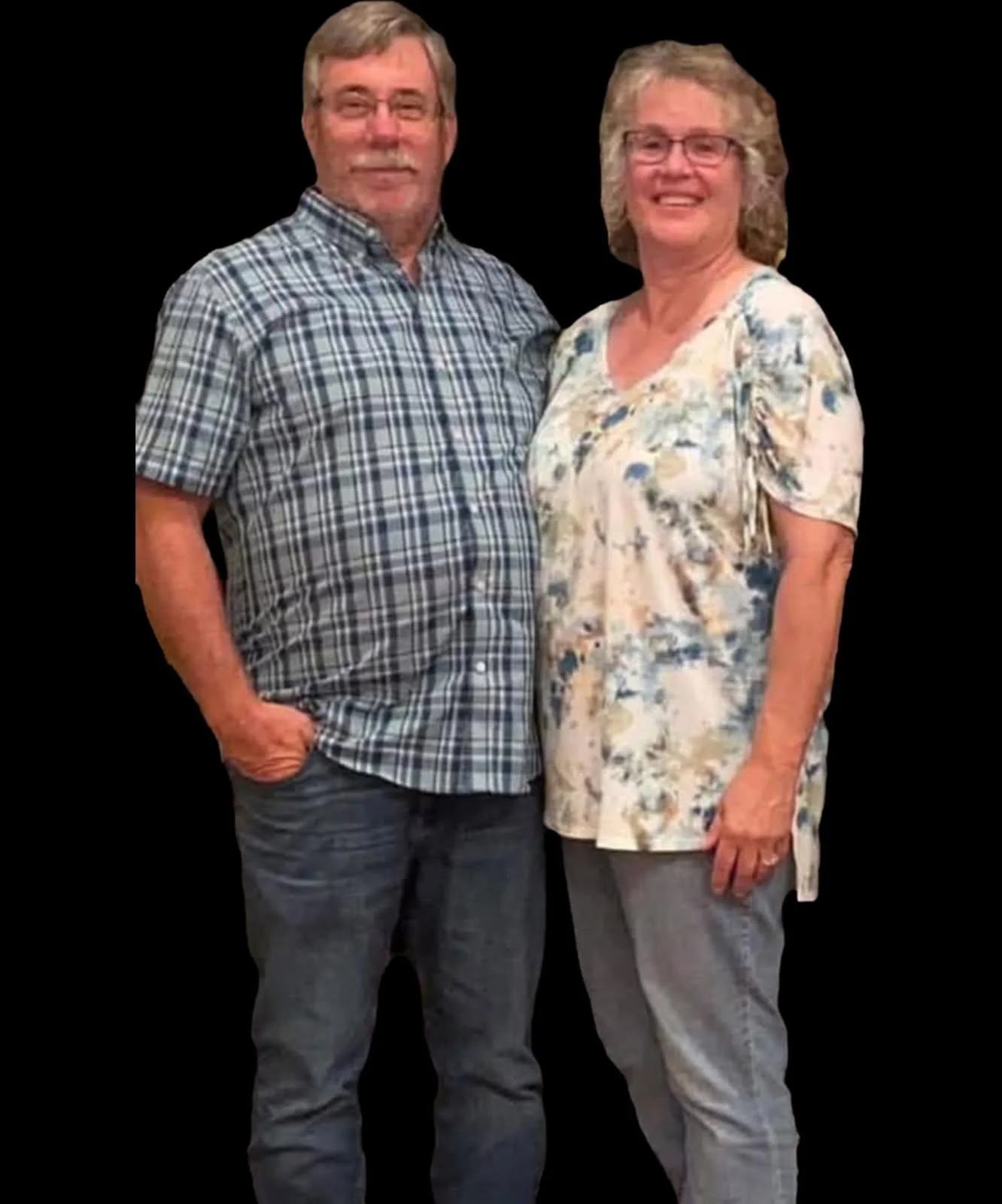 A middle-aged man and woman smiling and standing close together against a black background. The man is wearing a plaid shirt and jeans, and the woman is wearing a floral blouse and gray pants.