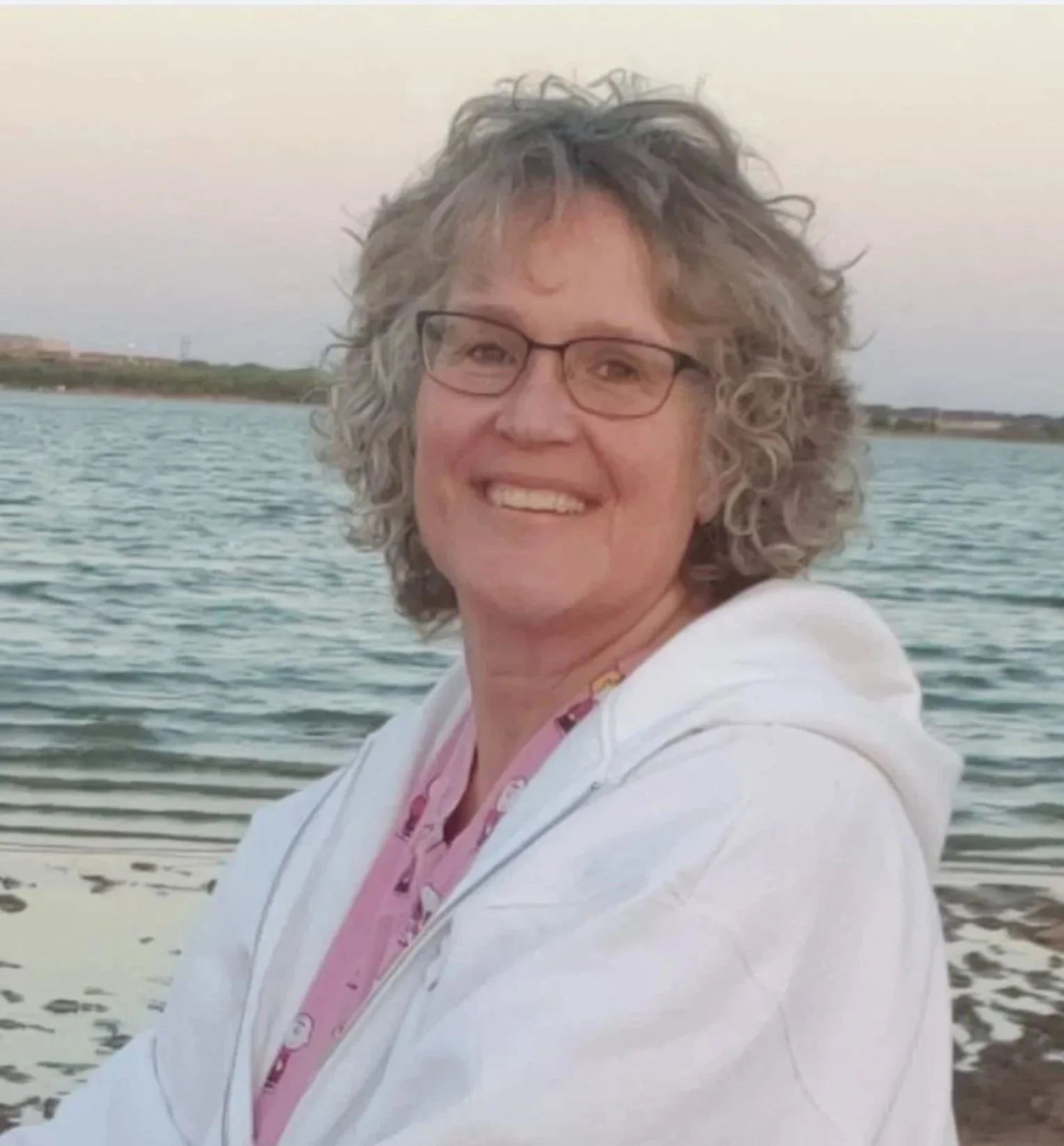A woman with curly gray hair and glasses smiling outdoors near a body of water, with land and sky in the background during sunset.
