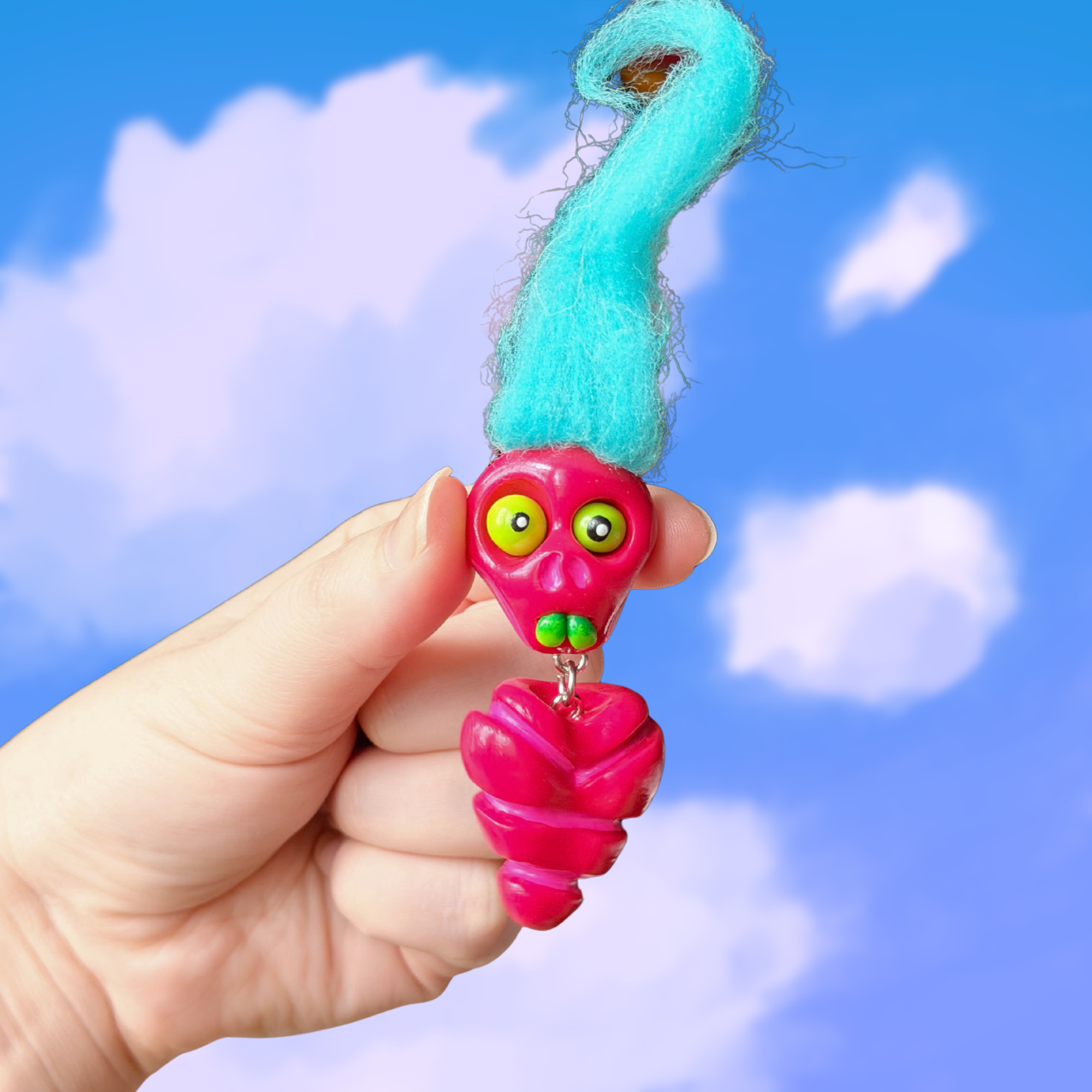 Polymer clay berry colored fidget that looks like a quirky skeleton with no arms or legs and blue hair. Photographed on top of a cloudy sky background.