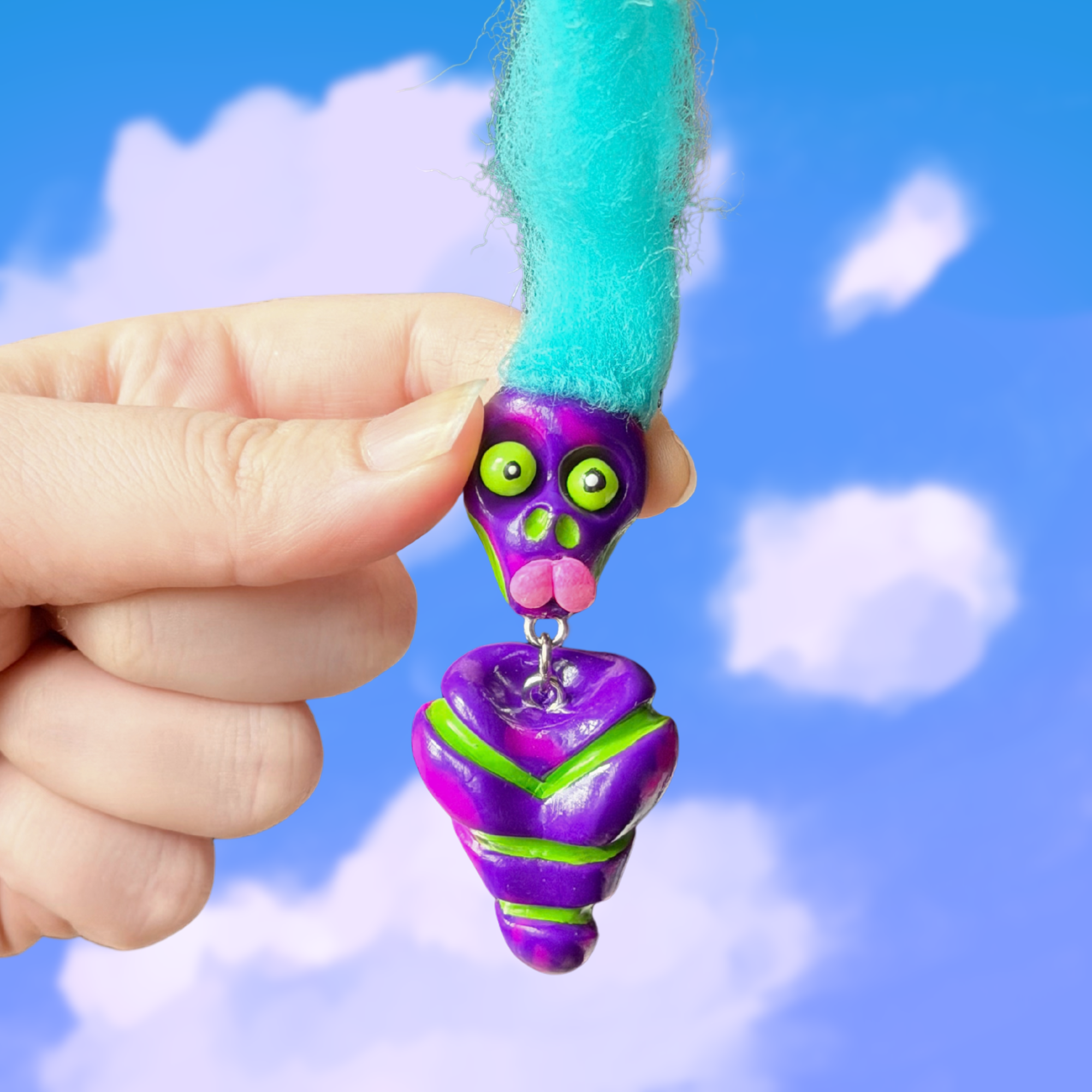 Polymer clay spotted purple colored fidget that looks like a quirky skeleton with no arms or legs and blue hair. Photographed on top of a cloudy sky background.