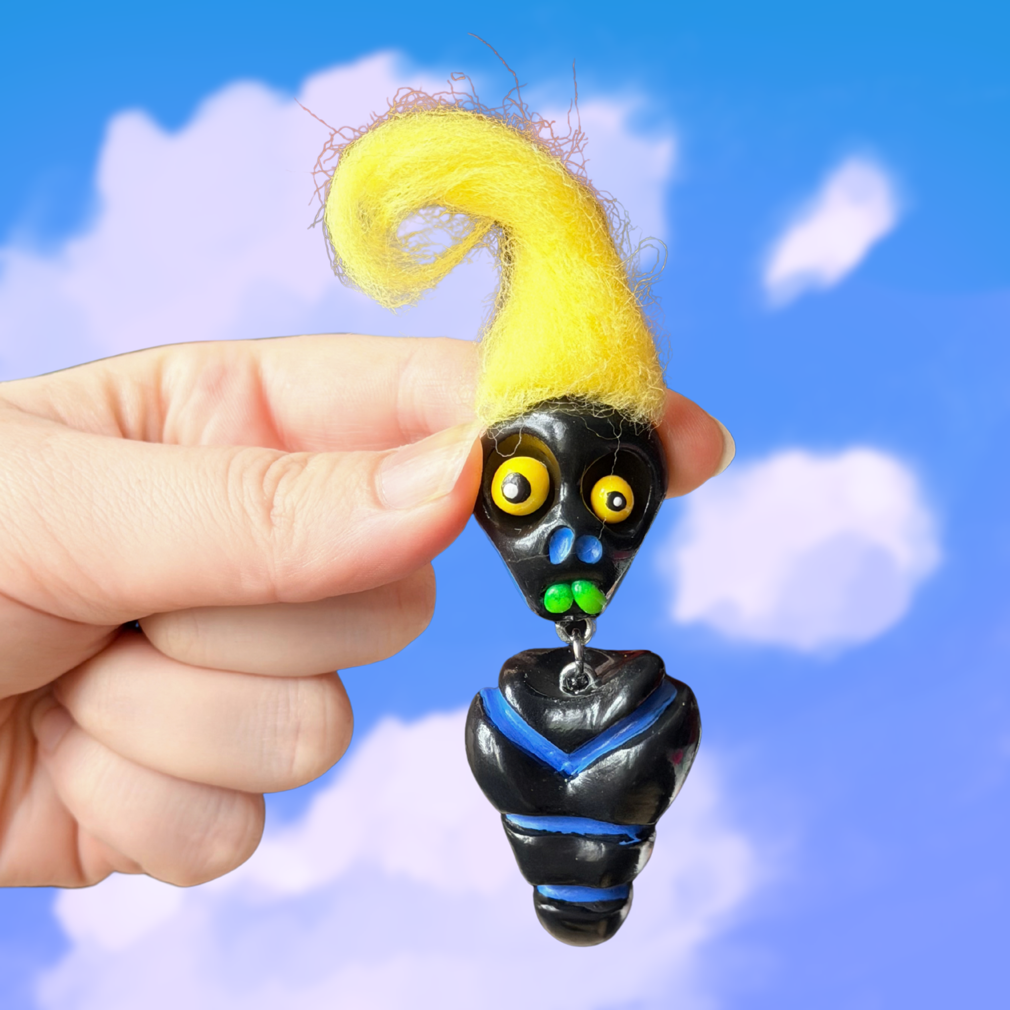 Polymer clay black colored fidget that looks like a quirky skeleton with no arms or legs and yellow hair. Photographed on top of a cloudy sky background.
